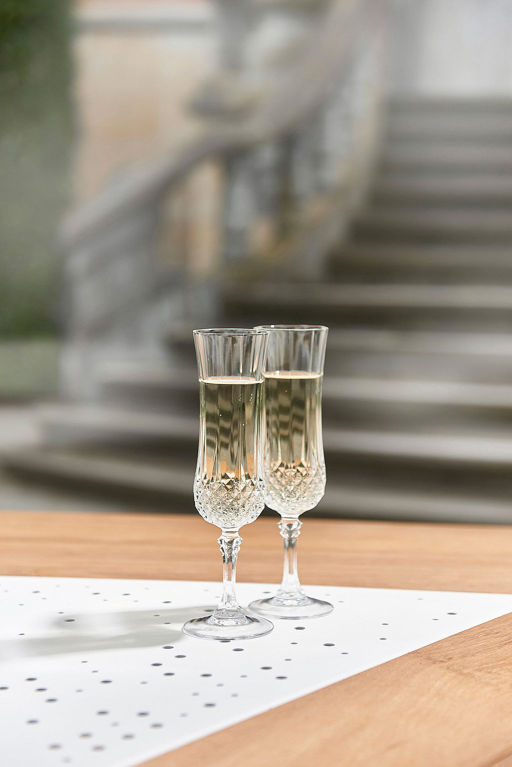 Deux coupes de champagne en cristal remplies de vin pétillant sont posées sur une table de jardin Anker en bois avec incrustations métalliques perforées, avec un escalier en pierre flou et un jardin en arrière-plan.