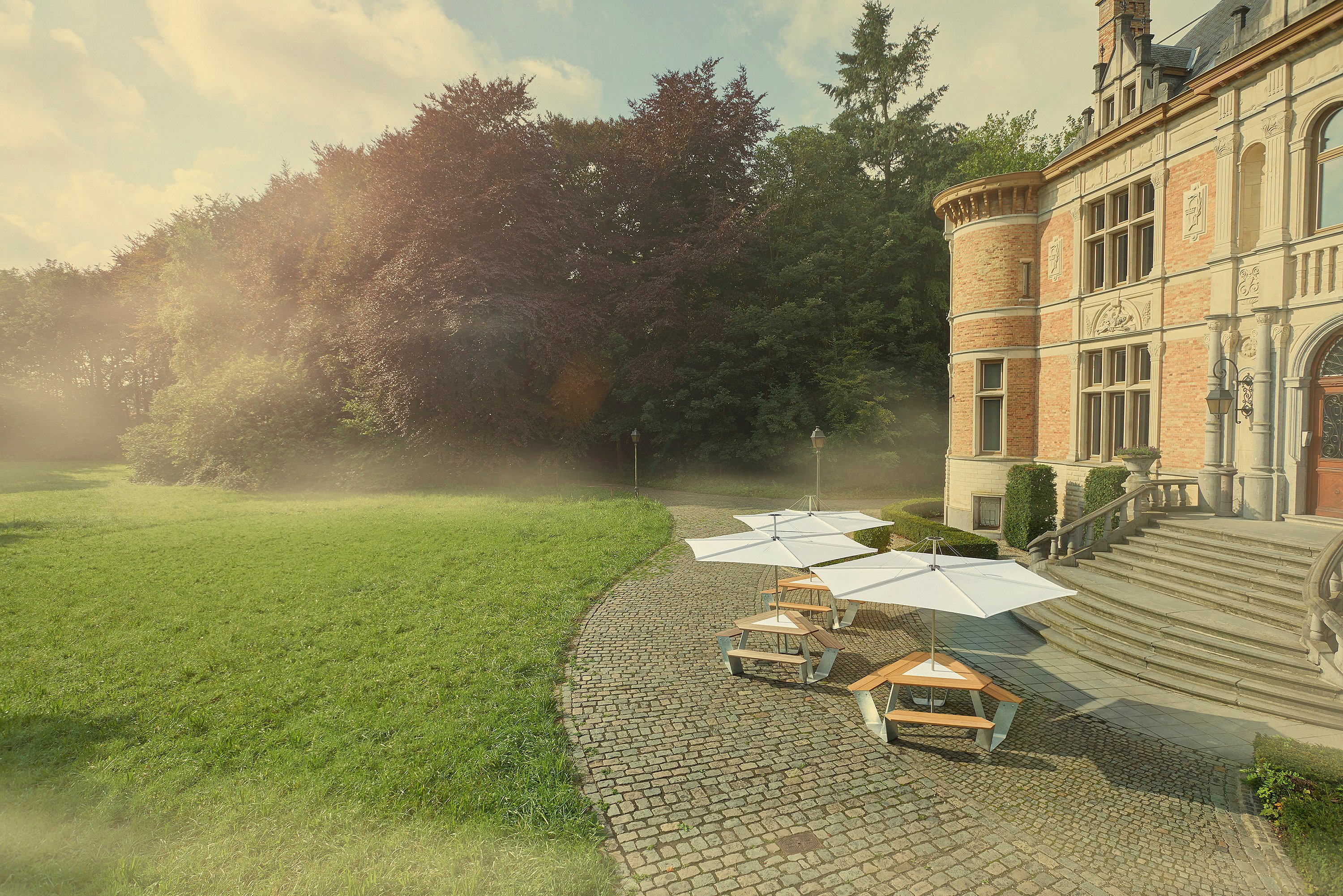   Espace salon extérieur avec tables triangulaires Anker sous de grands parasols blancs Inumbrina donnant sur les jardins paysagers d'un château historique.