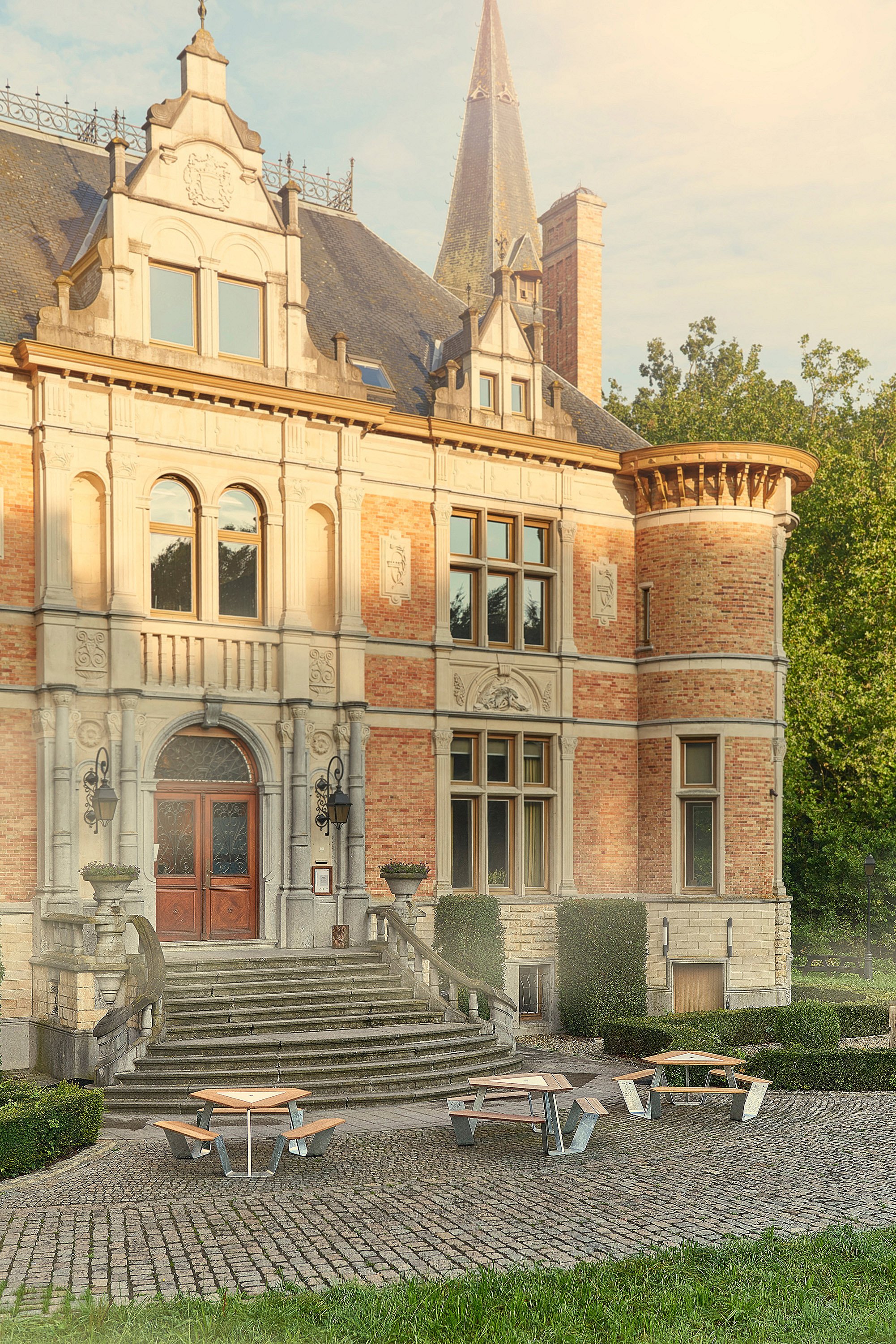   Espace salon extérieur avec tables triangulaires Anker sous de grands parasols blancs Inumbrina donnant sur les jardins paysagers d'un château historique.