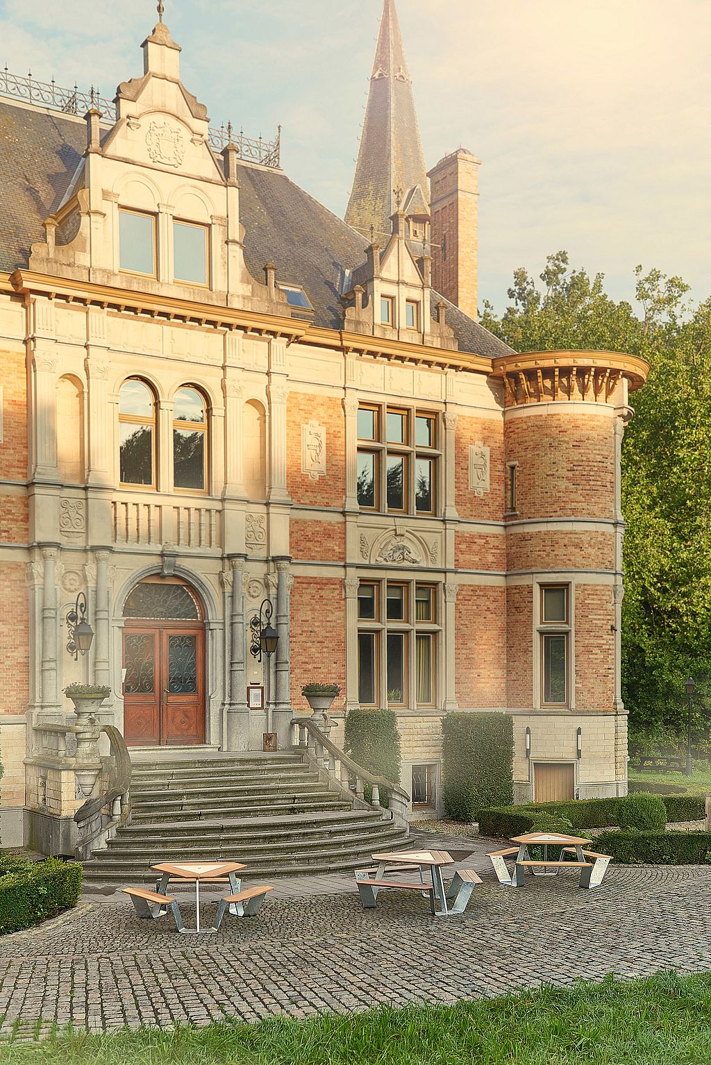 Espace salon extérieur avec tables triangulaires Anker sous de grands parasols blancs Inumbrina donnant sur les jardins paysagers d'un château historique.