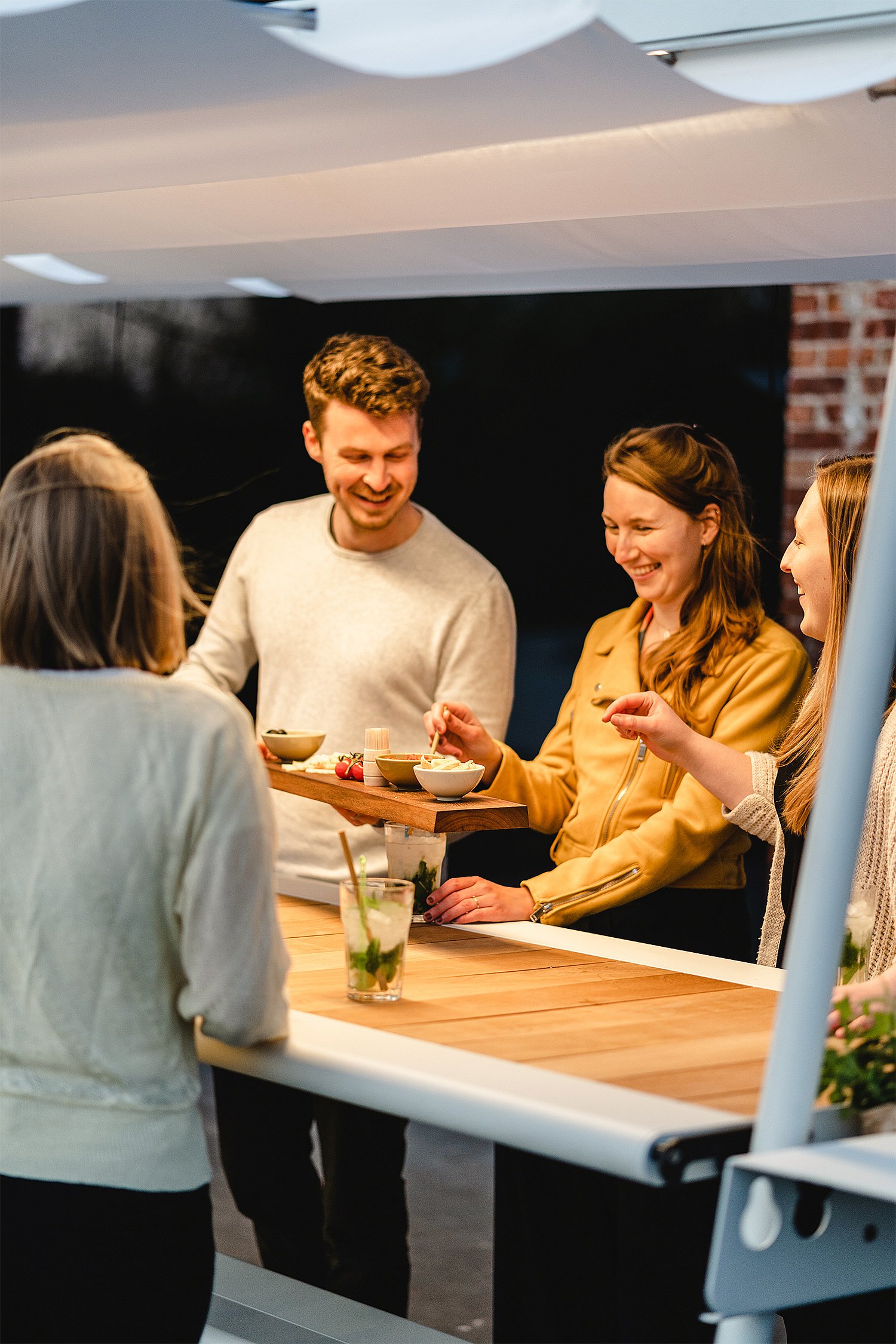 “Twee mensen toasten naast de AMAi buitentafel met vrienden verzameld in de schaduw. ”