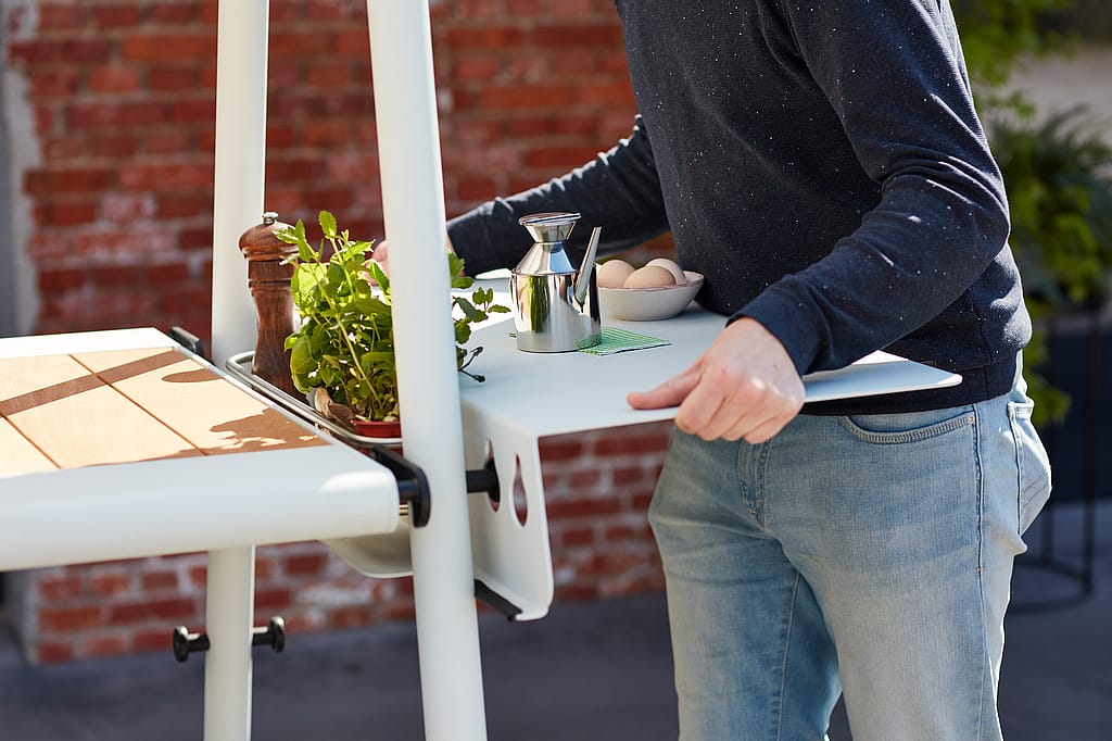 “Persoon die een zijschaal met kruiden en specerijen in het AMAi-tafelframe schuift terwijl hij buiten eten klaarmaakt. ”