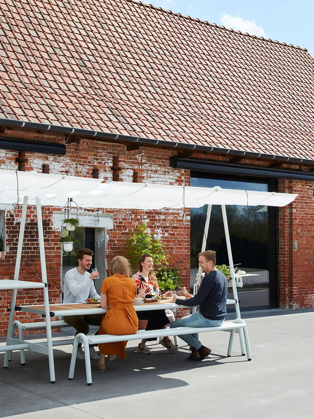 “Four people sitting and chatting around a long AMAi outdoor table with canopy in front of a rustic brick house, enjoying food together on a sunny day.”