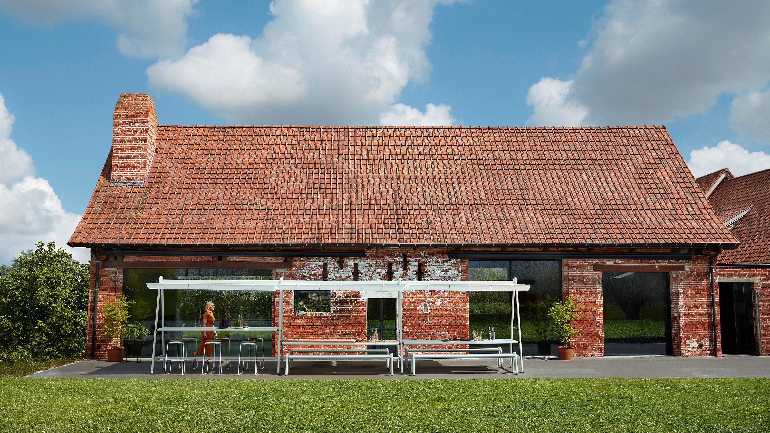 “Breed uitzicht op een bakstenen boerderij met een lange AMAi buitentafel onder een witte luifel op het terras, omgeven door een groen gazon. ”