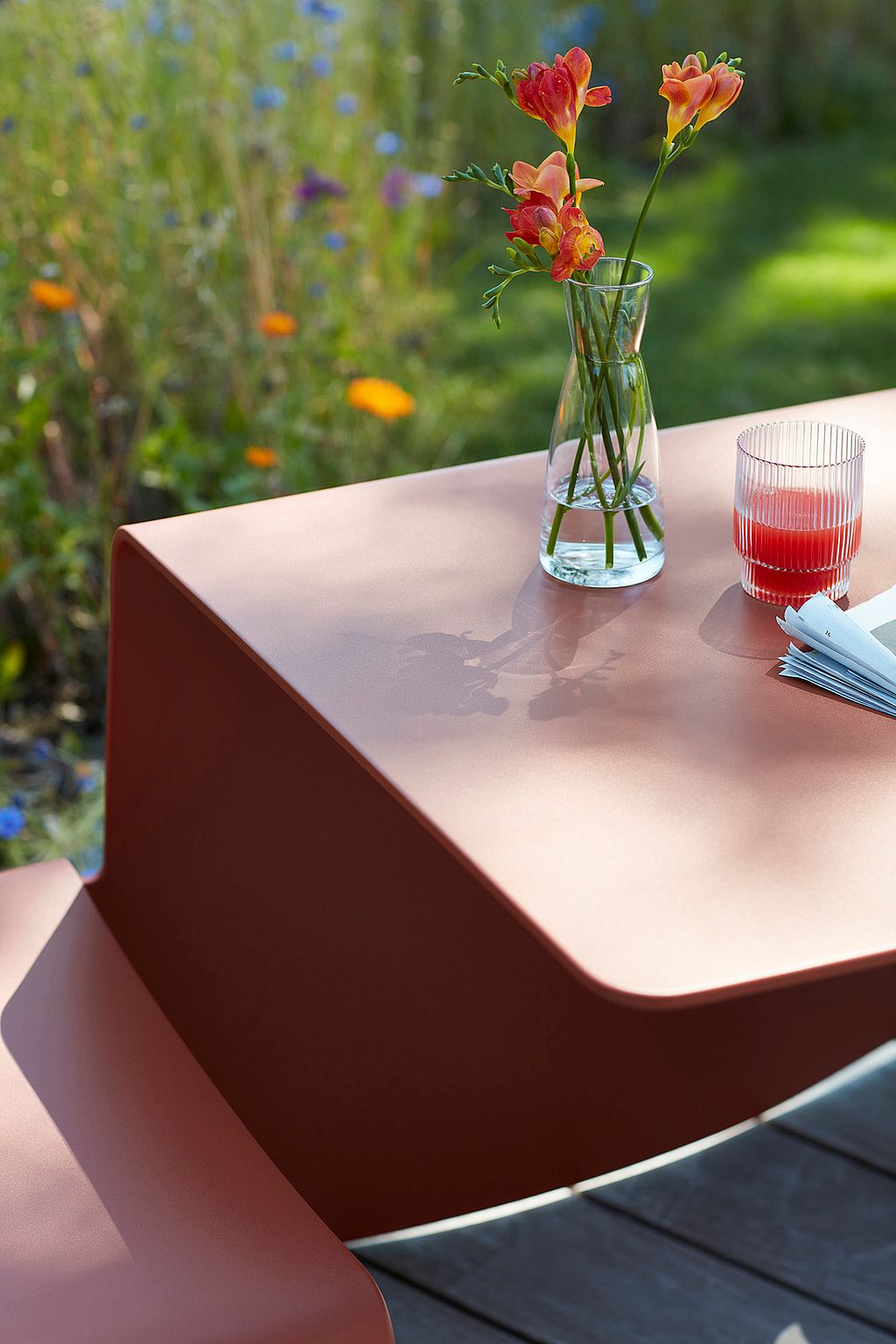 "Nahaufnahme der runden Ecke eines roten Terrakotta-Tisches Picnik mit einer orangefarbenen Glasvase und einem roten Getränk, auf einem verschwommenen Gartenhintergrund."