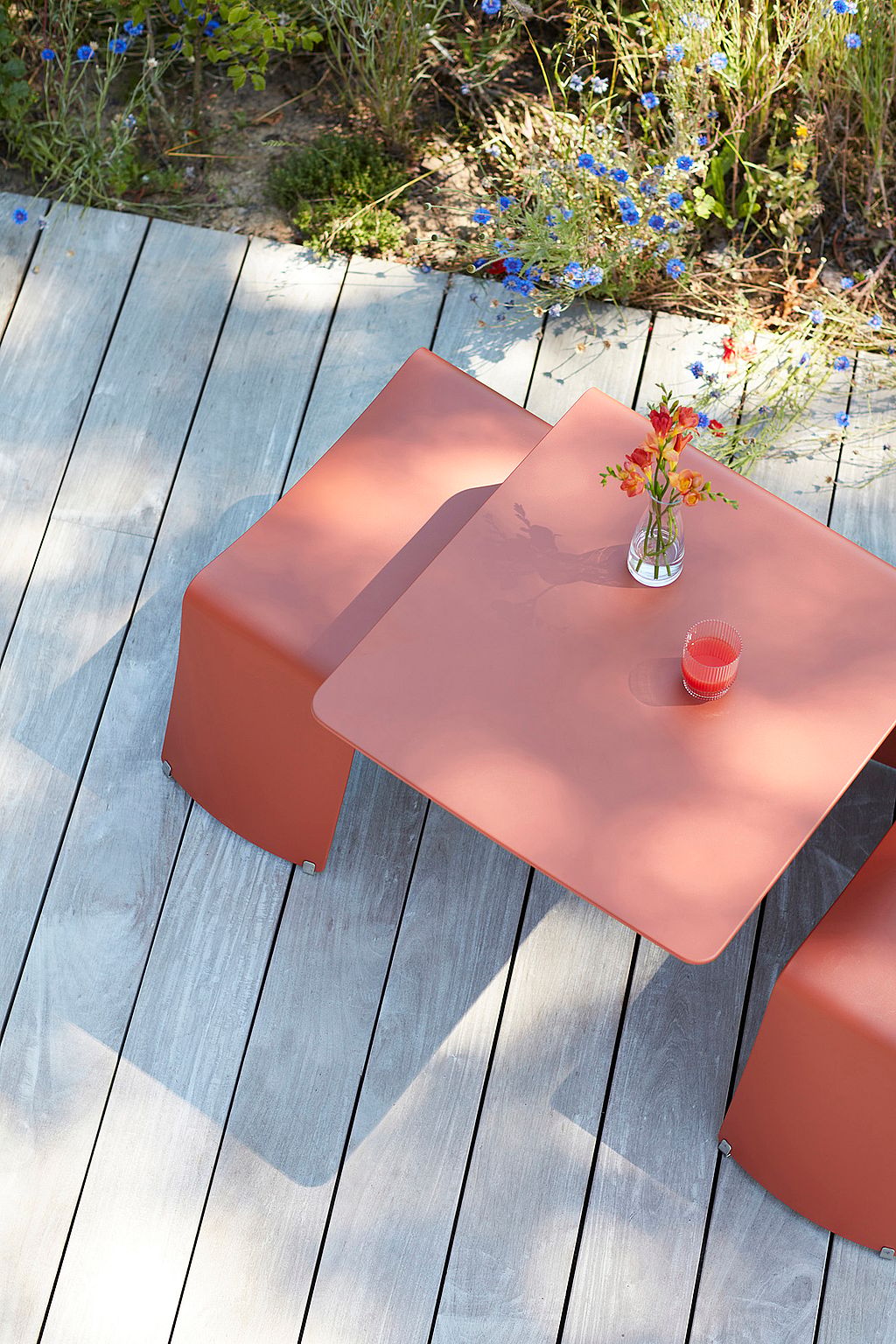 "Blick von oben auf einen roten Terrakotta-Picnik-Tisch und -Bänken auf einer Holzterrasse, mit einem Glas rotem Getränk und einer kleinen Vase mit Blumen über dem Tisch."