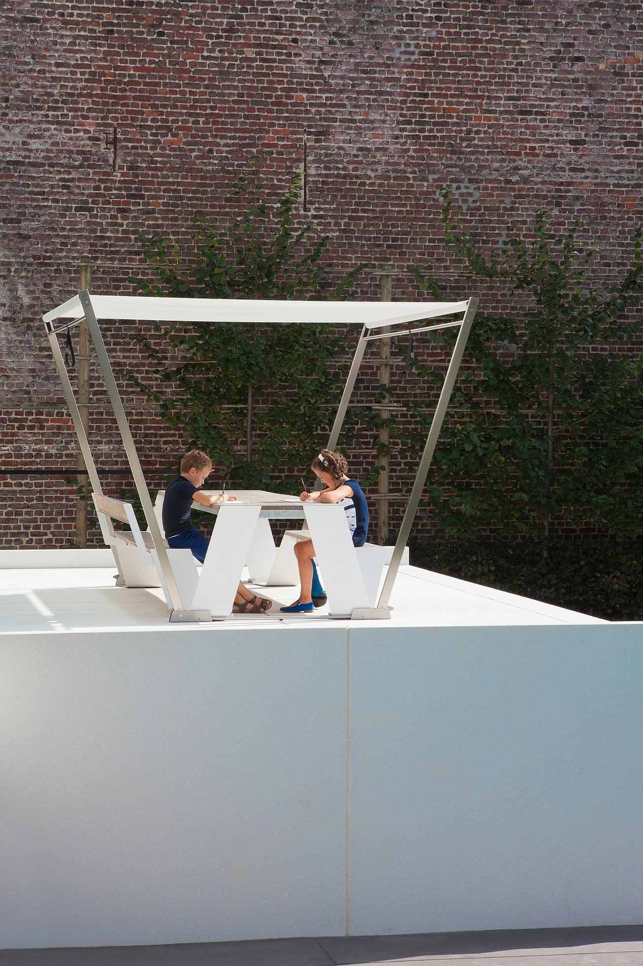 “Twee kinderen zaten tegenover elkaar aan een wit gearceerde picknicktafel op een minimalistisch terras, met een bakstenen muur erachter. ”