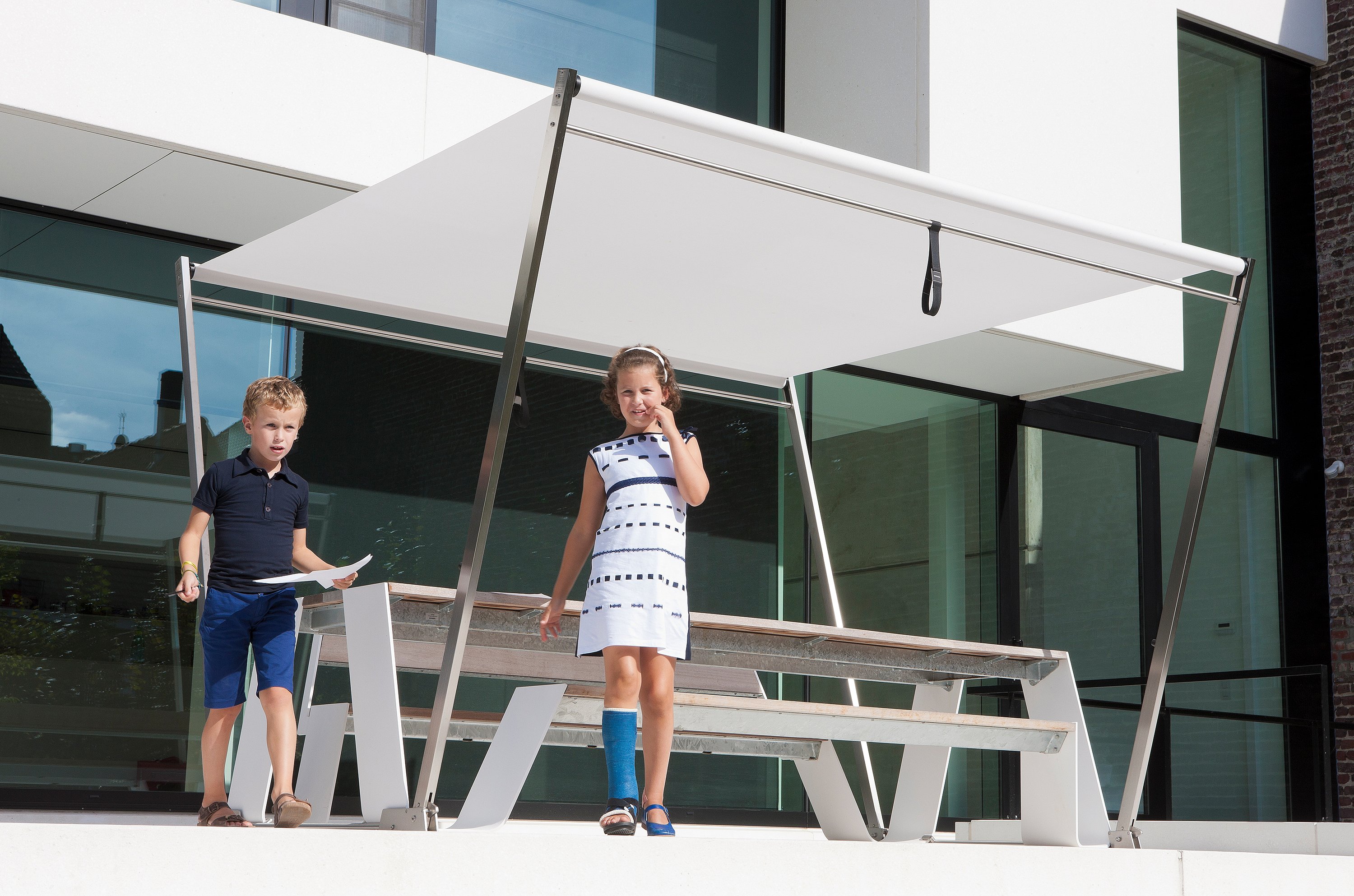 “Twee kinderen lopen langs een witte picknicktafel met een geïntegreerde parasol op een modern terras, met een glazen huis erachter. ”