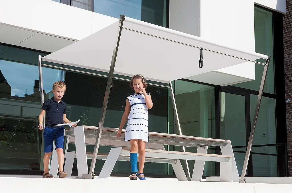 “Two children walking past a white picnic table with an integrated sunshade on a modern terrace, with a glass-fronted house behind them.”