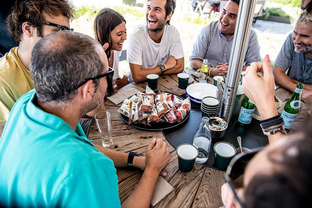 “Round picnic-style restaurant table set for guests on a sunny terrace, designed to encourage easy conversation.”