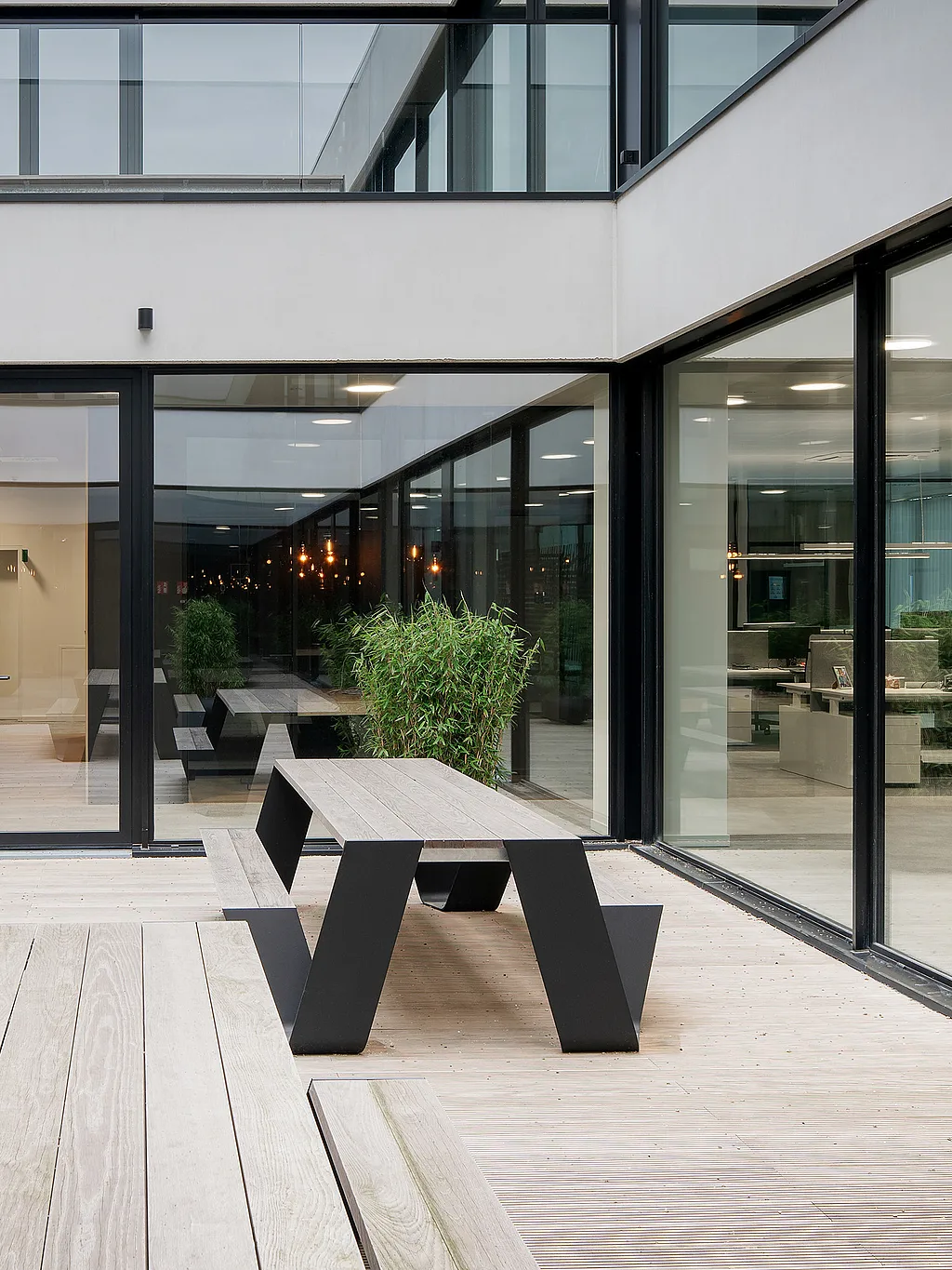 Modernes Bürointerieur mit bodentiefen Fenstern und einem offenen Terrassenbereich mit Hopper-Picknicktischen aus Holz, an denen Mitarbeiter im Freien arbeiten können.