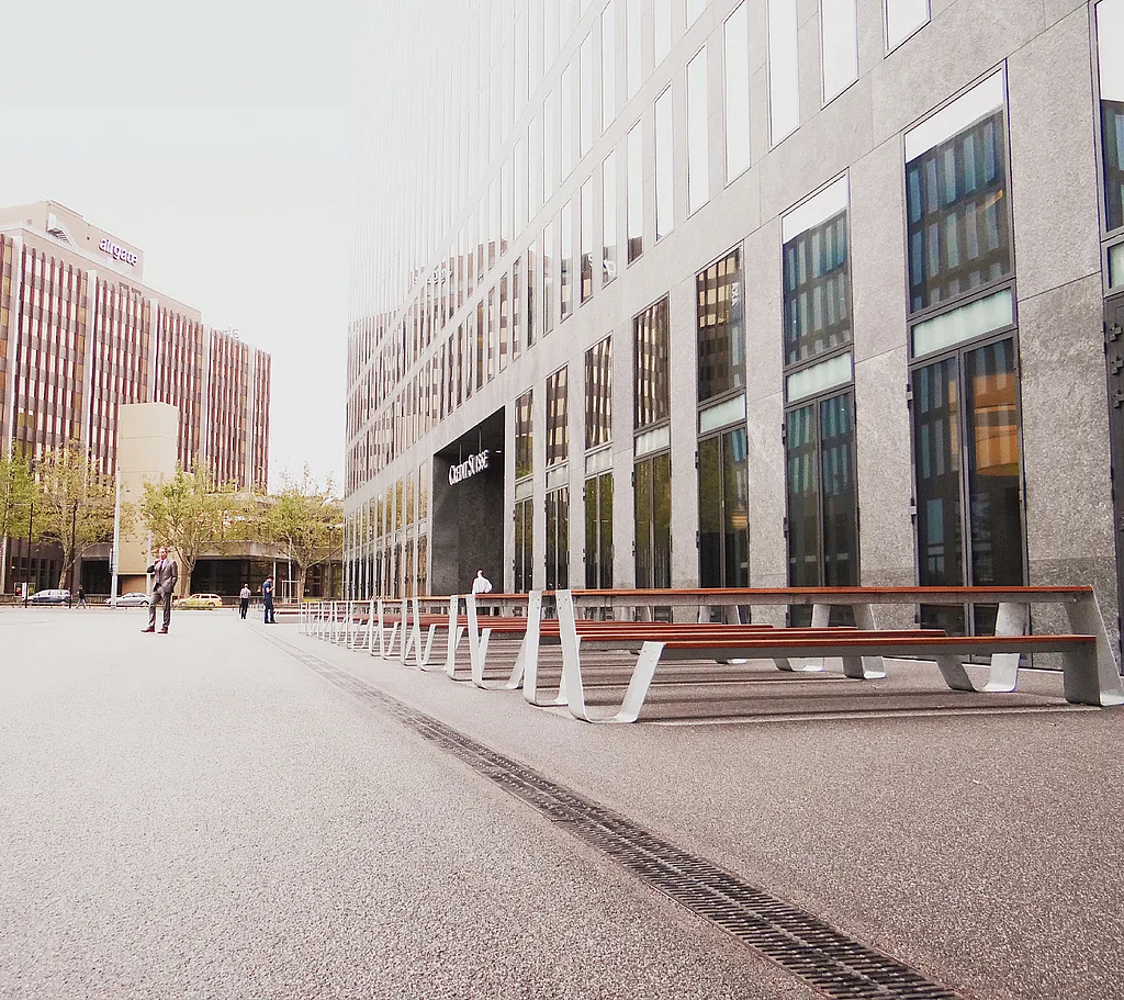 “Long row of white Hopper picnic tables lined up in an urban plaza beside the Credit Suisse tower in Zürich, with the glass-and-steel high-rise rising in the background.”