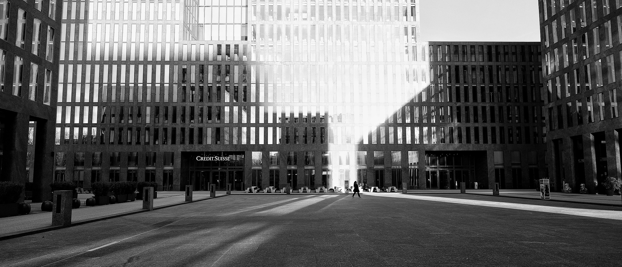 « Plan extérieur large de l’esplanade de la tour du Credit Suisse, où une ligne répétée de tables Hopper structure l’espace public entre des bâtiments contemporains. »