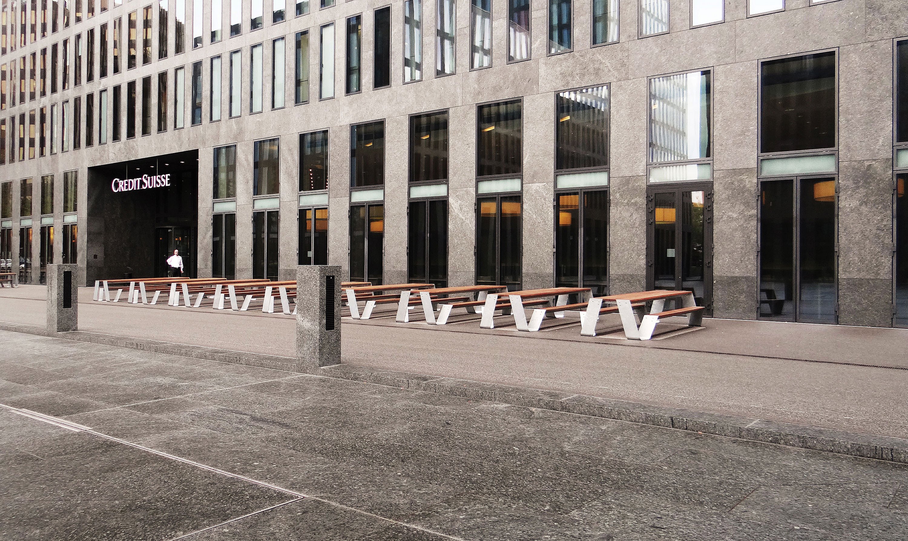 « Place publique avec plusieurs tables de pique-nique Hopper blanches disposées en ligne droite, créant des sièges généreux devant les tours de bureaux modernes. »