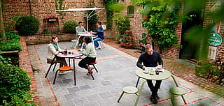 “Guests tell stories about virus tables in a hidden courtyard shaded by an acacia roof. ”