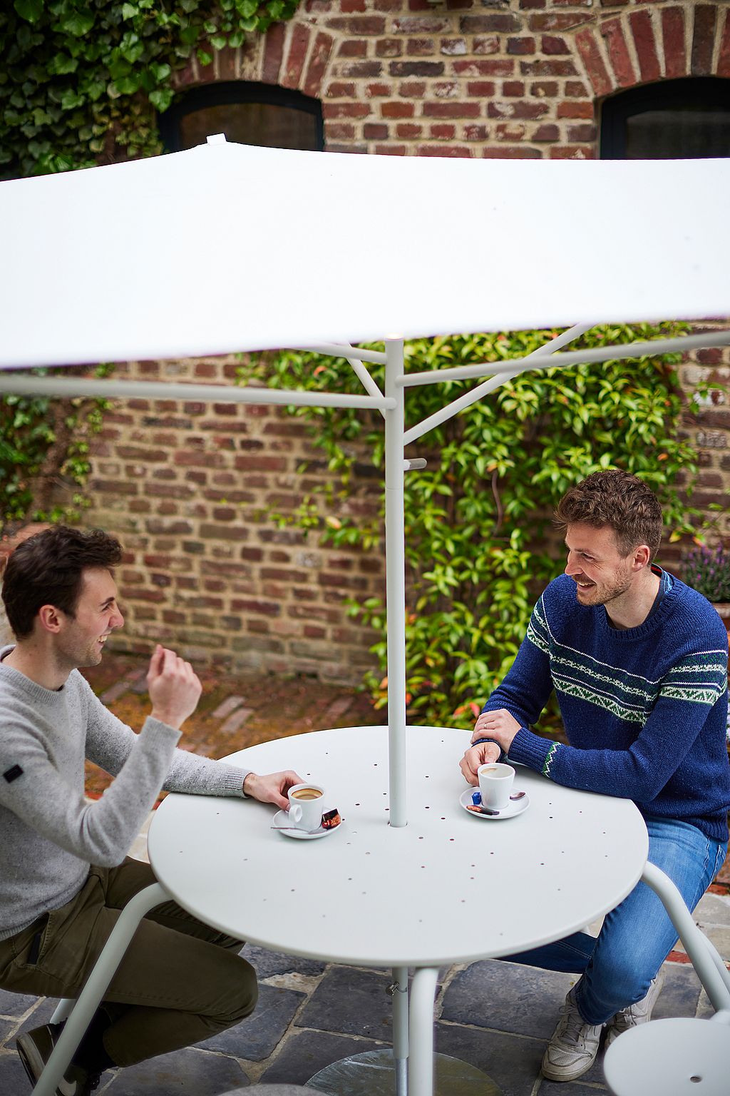 “Guests tell stories about virus tables in a hidden courtyard shaded by an acacia roof. ”