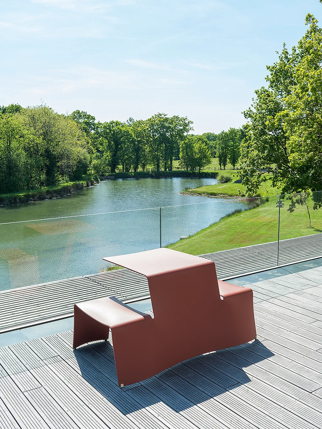 “Roter Picnik-Tisch auf einer Dachterrasse mit Blick auf einen ruhigen Teich, mit Glasbalustraden, Bäumen und offenem Himmel im Hintergrund.”