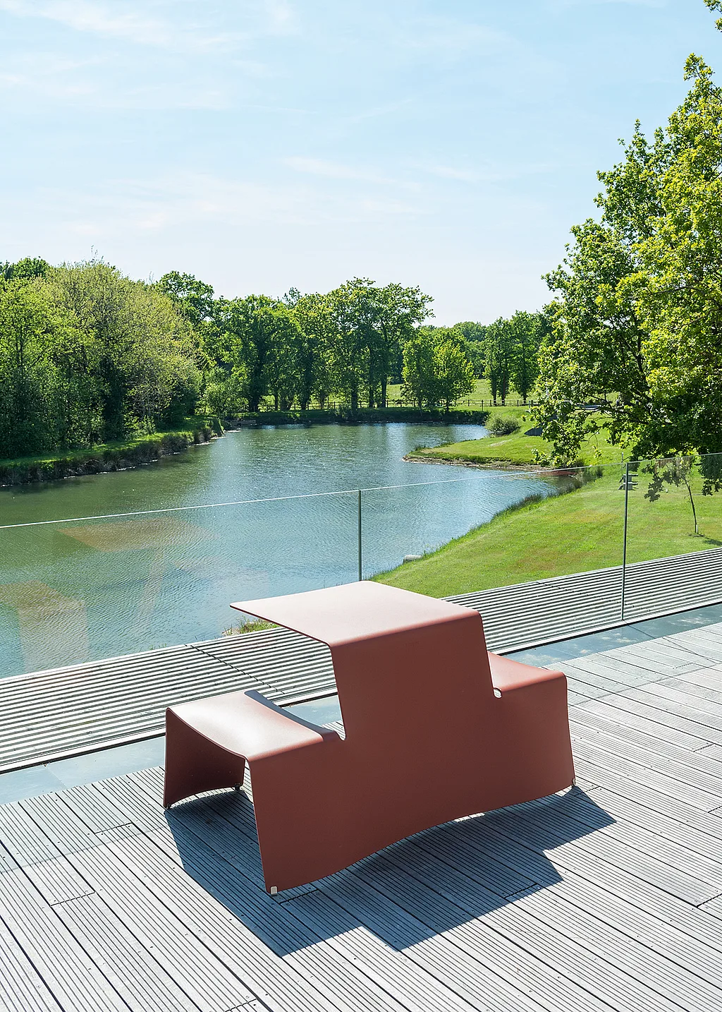« Table Red Picnik sur une terrasse sur le toit donnant sur un étang calme, avec des balustrades en verre, des arbres et un ciel ouvert en arrière-plan. »