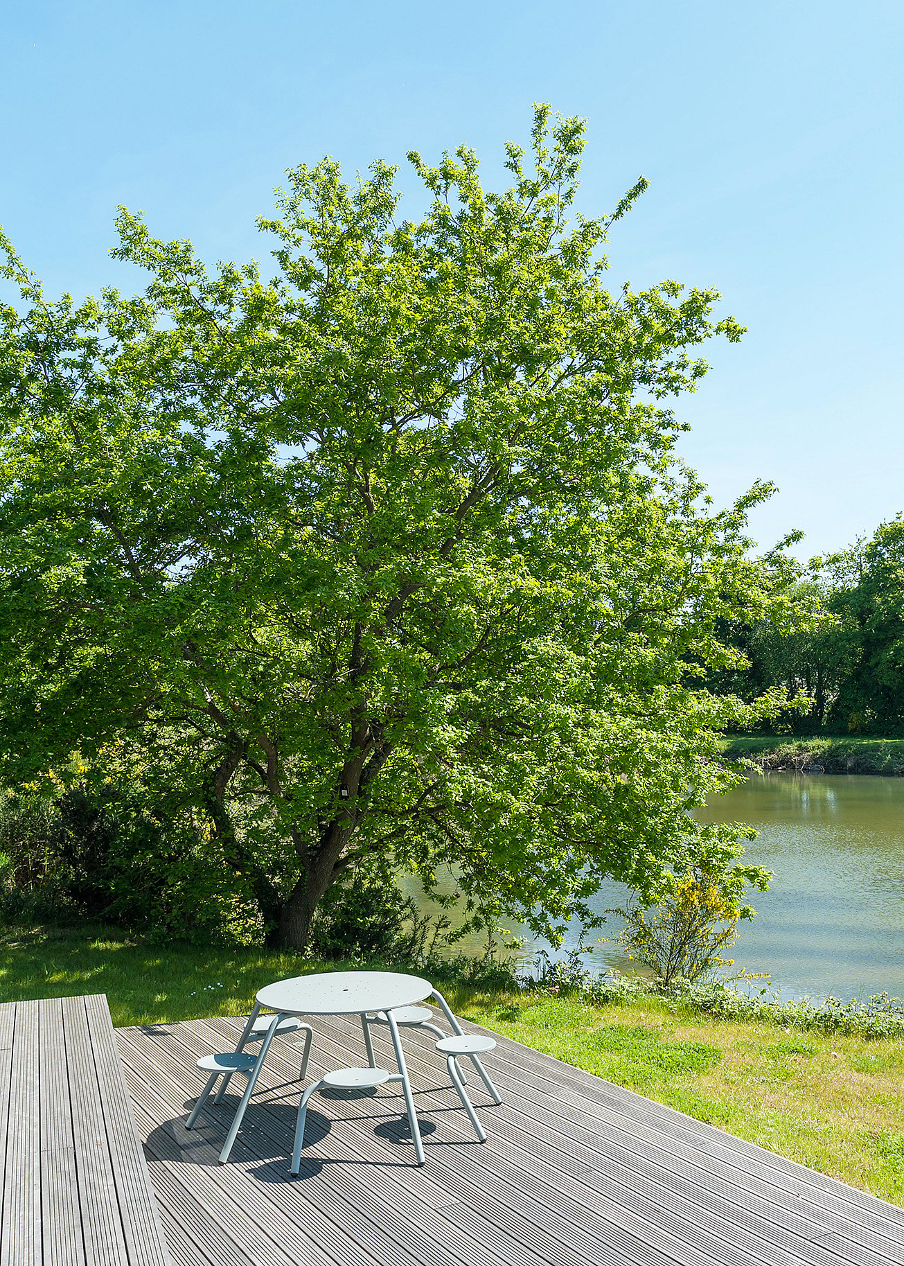 Enkele lichtgrijze ronde Virus tafel met bevestigde krukken geplaatst op een houten dek onder een grote bladboom naast het water.