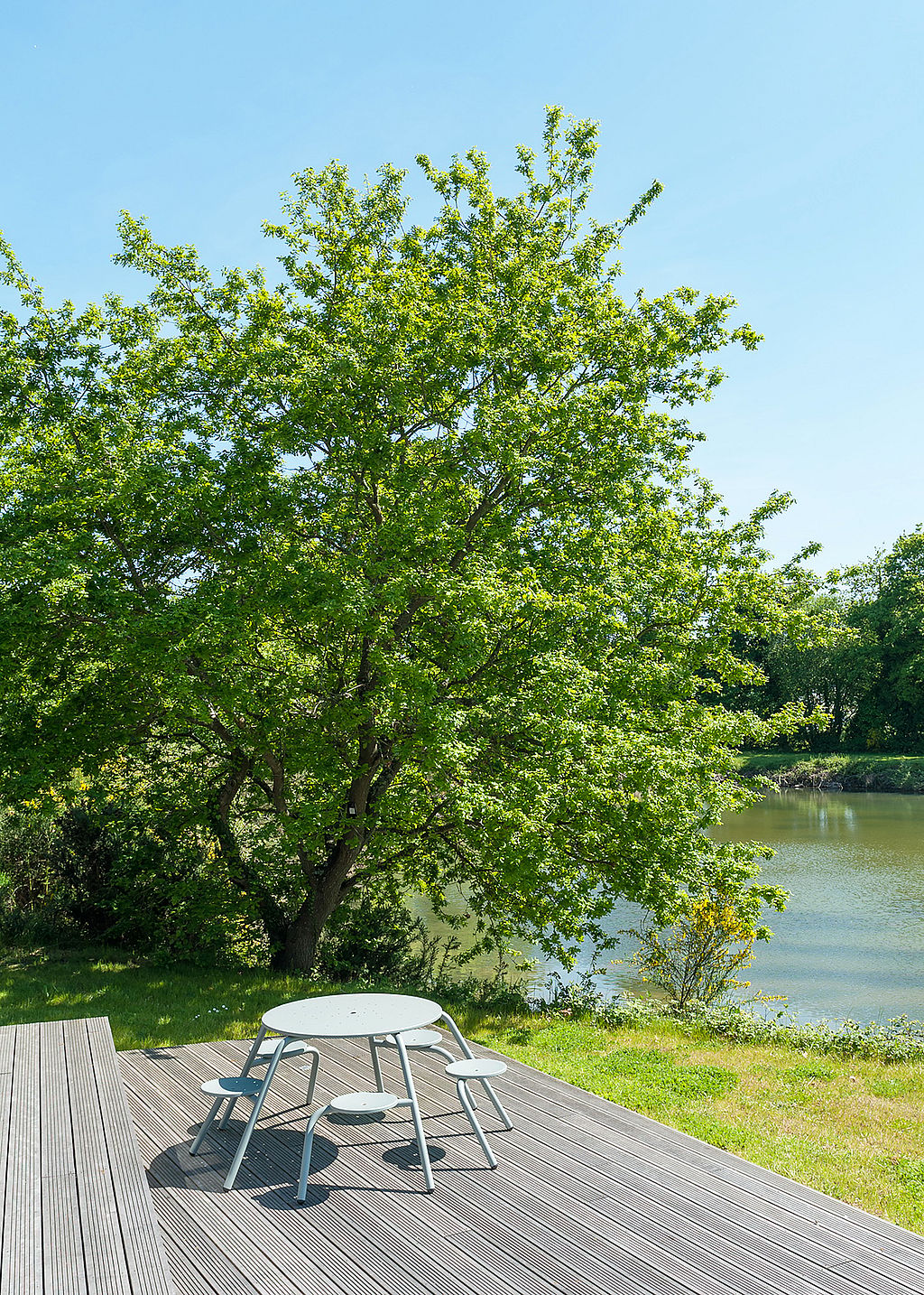 Enkele lichtgrijze ronde Virus tafel met bevestigde krukken geplaatst op een houten dek onder een grote bladboom naast het water.