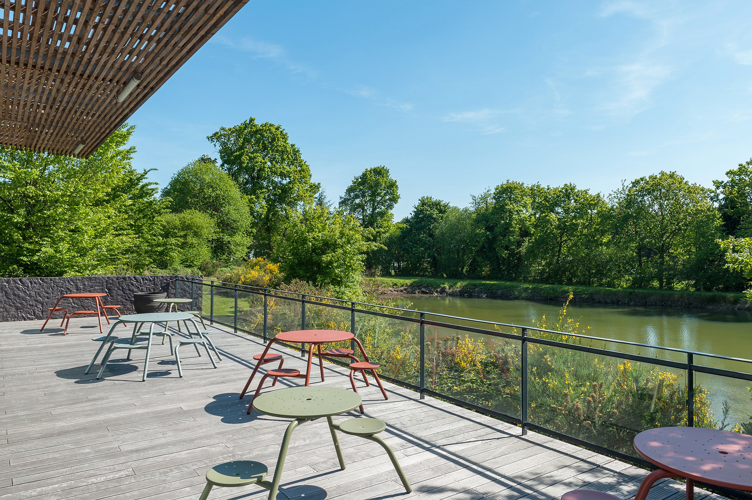 “Bovenaanzicht van twee kleurrijke Virustafels op een lager terras naast de vijver, omlijst door de glazen gevel van het gebouw en gelaagde daklijnen. ”