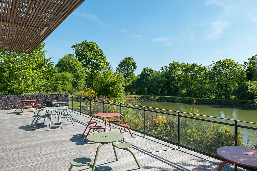 “Bovenaanzicht van twee kleurrijke Virustafels op een lager terras naast de vijver, omlijst door de glazen gevel van het gebouw en gelaagde daklijnen. ”