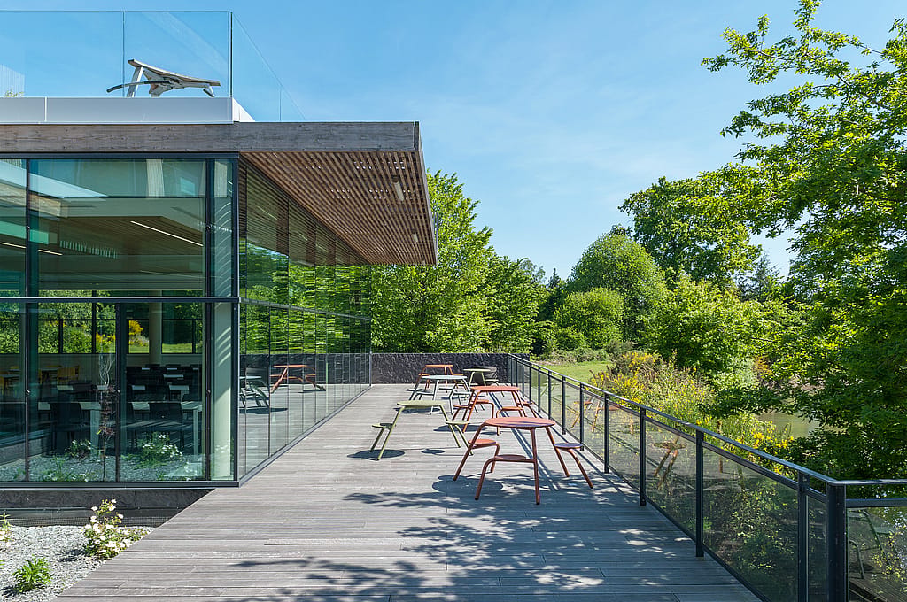 “Terras bekleed met kleurrijke ronde Virustafels en geïntegreerde krukken, naast een glazen reling met uitzicht over de vijver en het omliggende groen. ”
