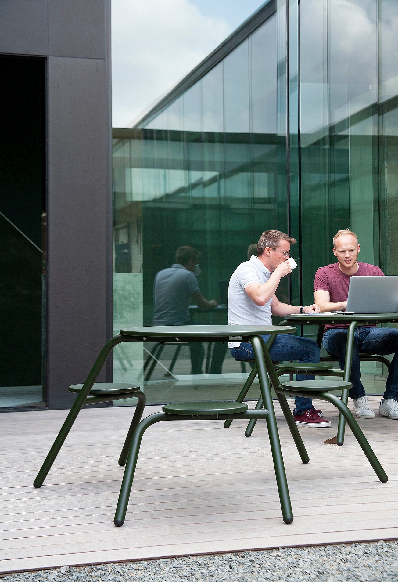 Two colleagues sit side by side at a green Virus table, one drinking from a cup while the other looks at a laptop, with the courtyard’s glass wall reflecting the scene behind them.