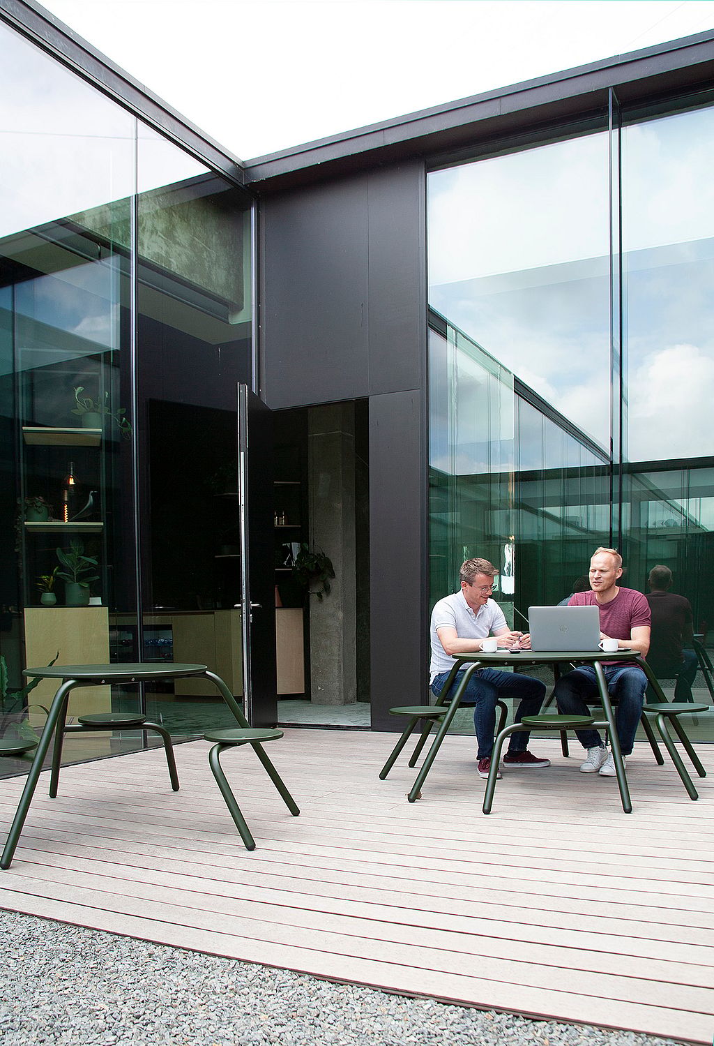 Two men brainstorm at a compact green Virus table in a modern glass courtyard, surrounded by reflective façades, an open doorway, and additional matching tables nearby.