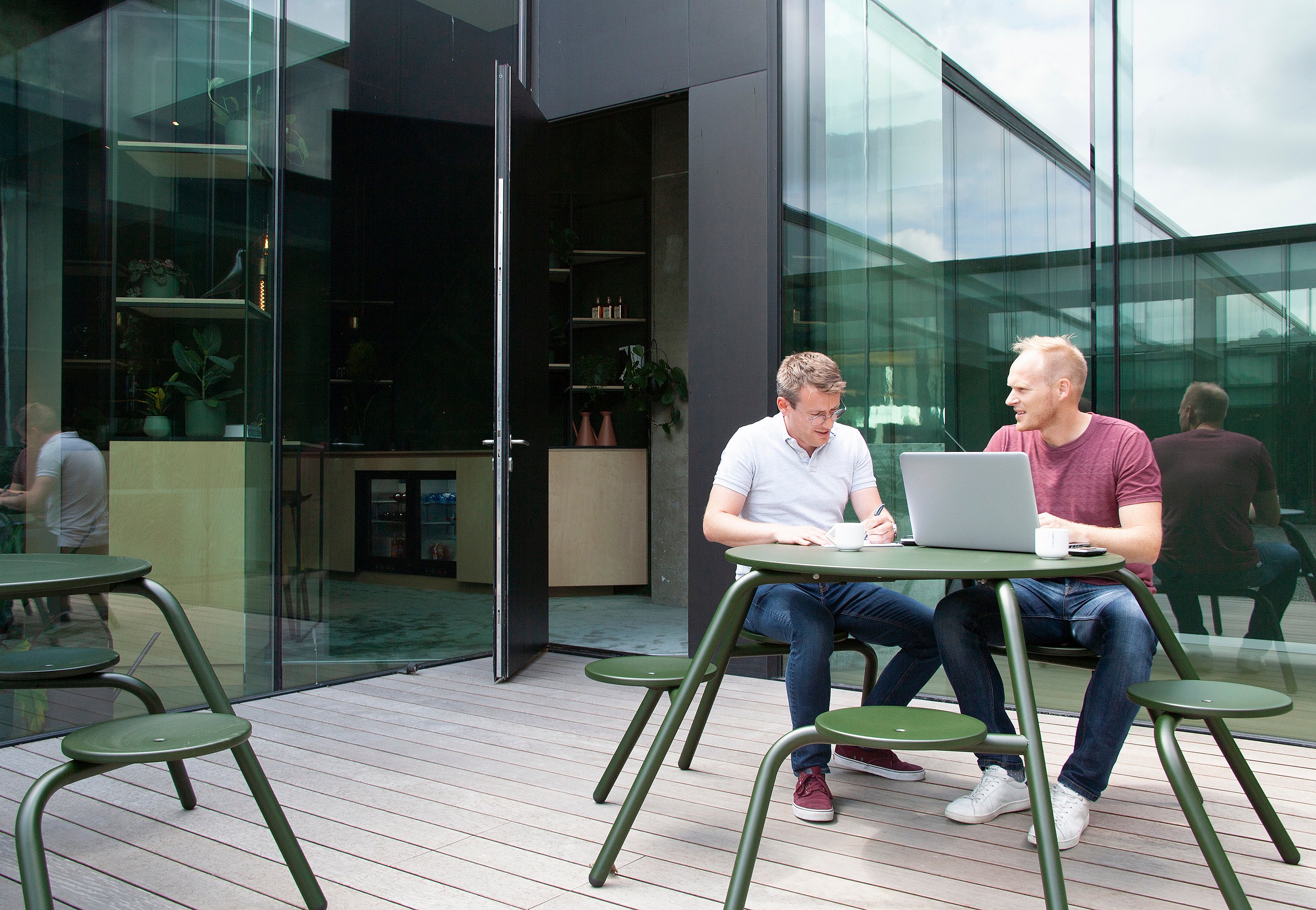 “Twee collega's zitten aan een donkergroene ronde Virus-tafel op een houten dek in een glazen wand binnenplaats en werken samen over een laptop met koffiekopjes op het tafelblad. ”
