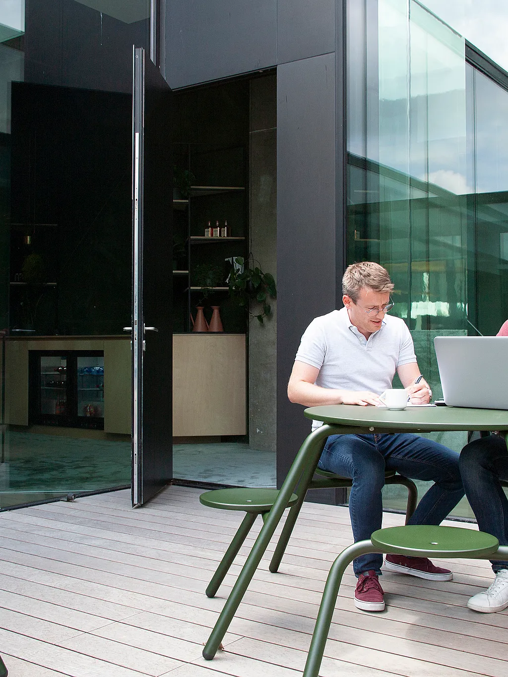 “Twee collega's zitten aan een donkergroene ronde Virus-tafel op een houten dek in een glazen wand binnenplaats en werken samen over een laptop met koffiekopjes op het tafelblad. ”