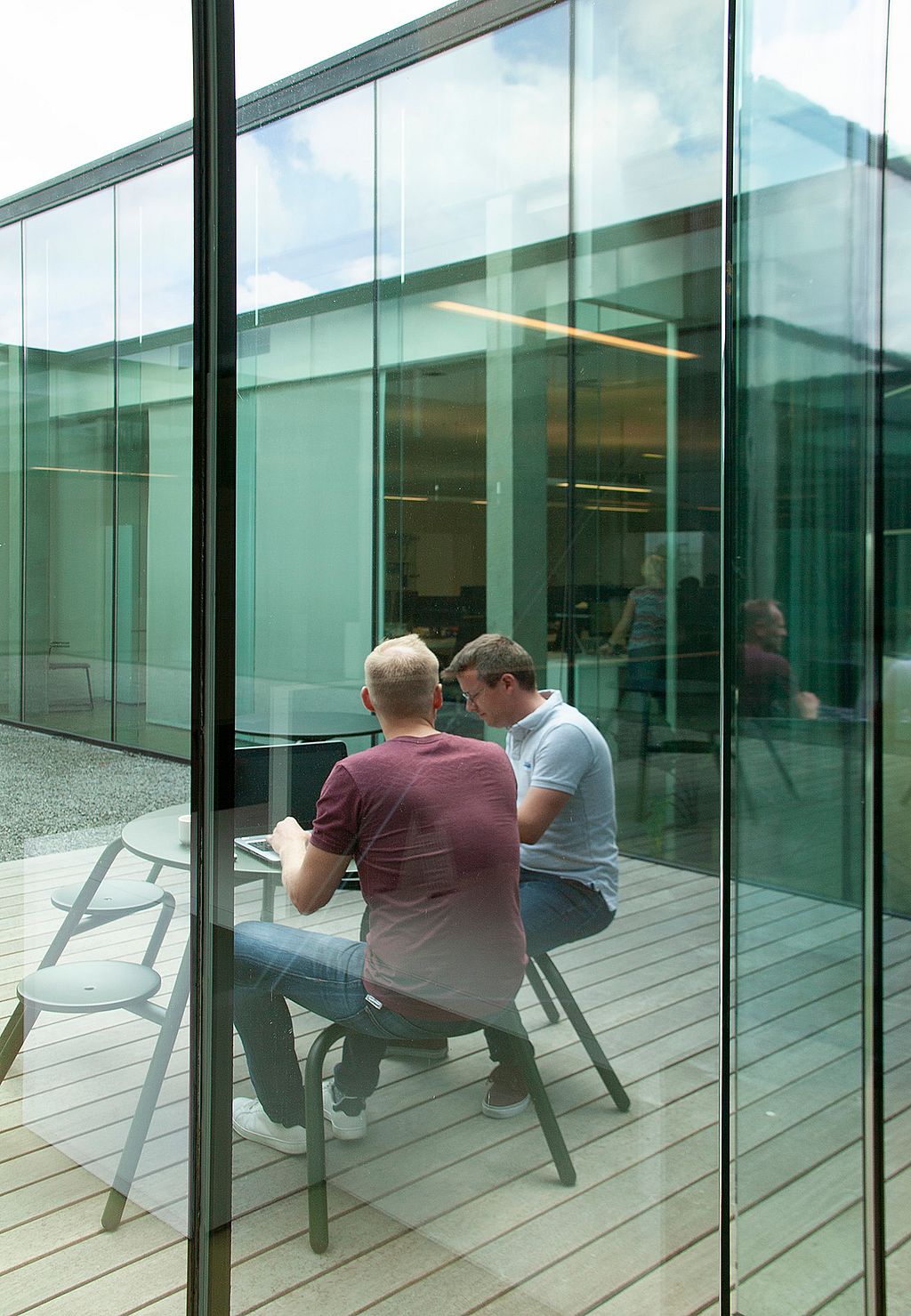 Viewed through tall glass panels, two colleagues sit in conversation at a green Virus table on the courtyard deck, with more matching tables in the foreground.