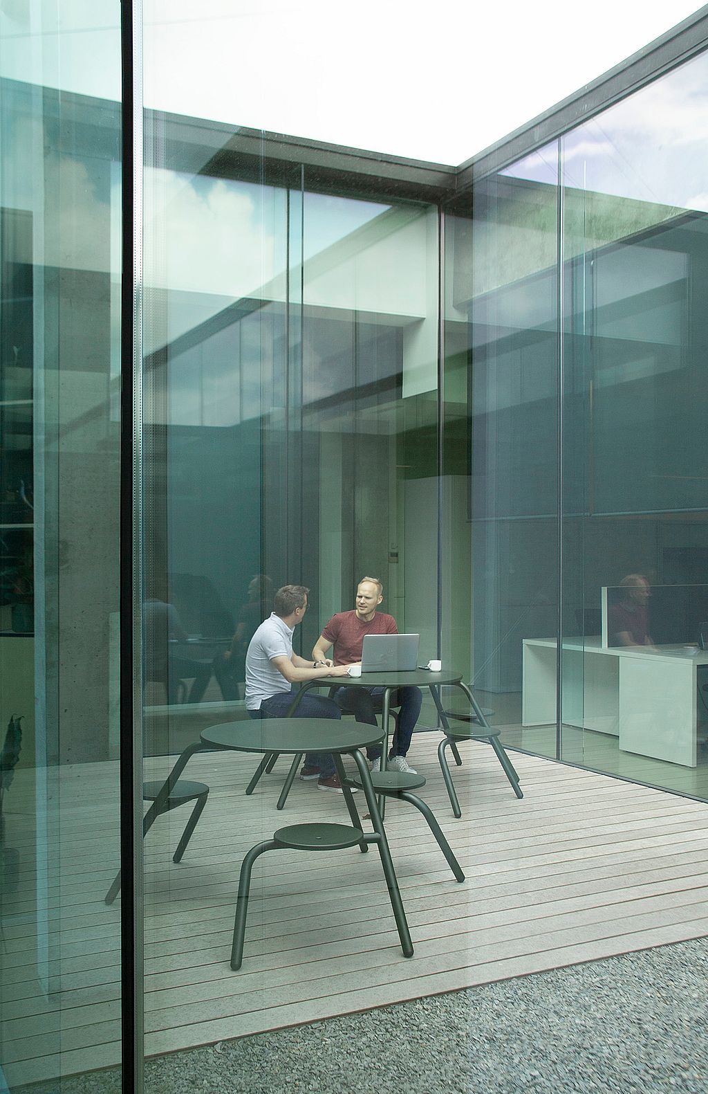 Two men brainstorm at a compact green Virus table in a modern glass courtyard, surrounded by reflective façades, an open doorway, and additional matching tables nearby.