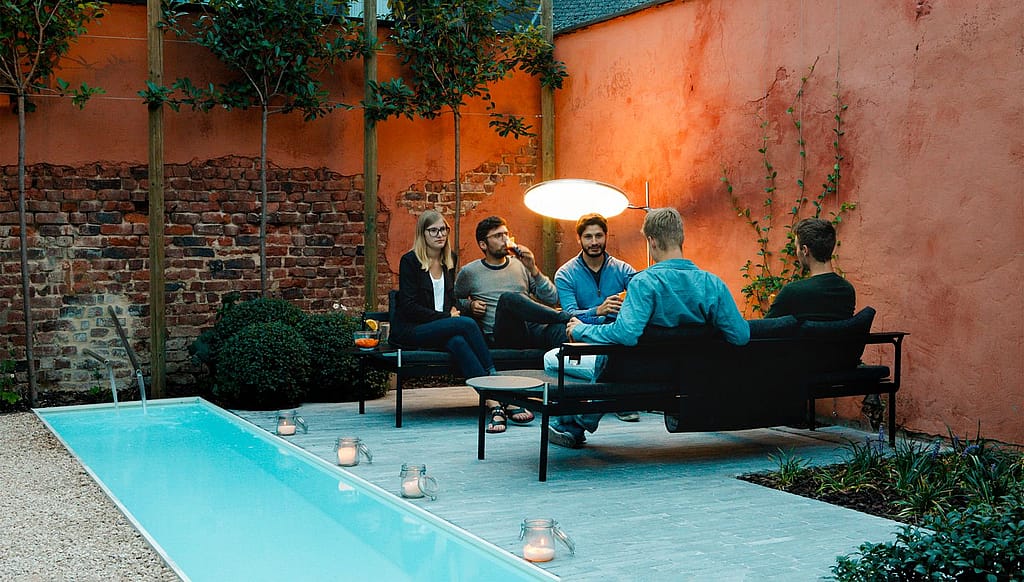 Fünf Freunde sitzen in der Abenddämmerung auf schwarzen Gartenliegen am beleuchteten Pool in einem gemütlichen Innenhof.