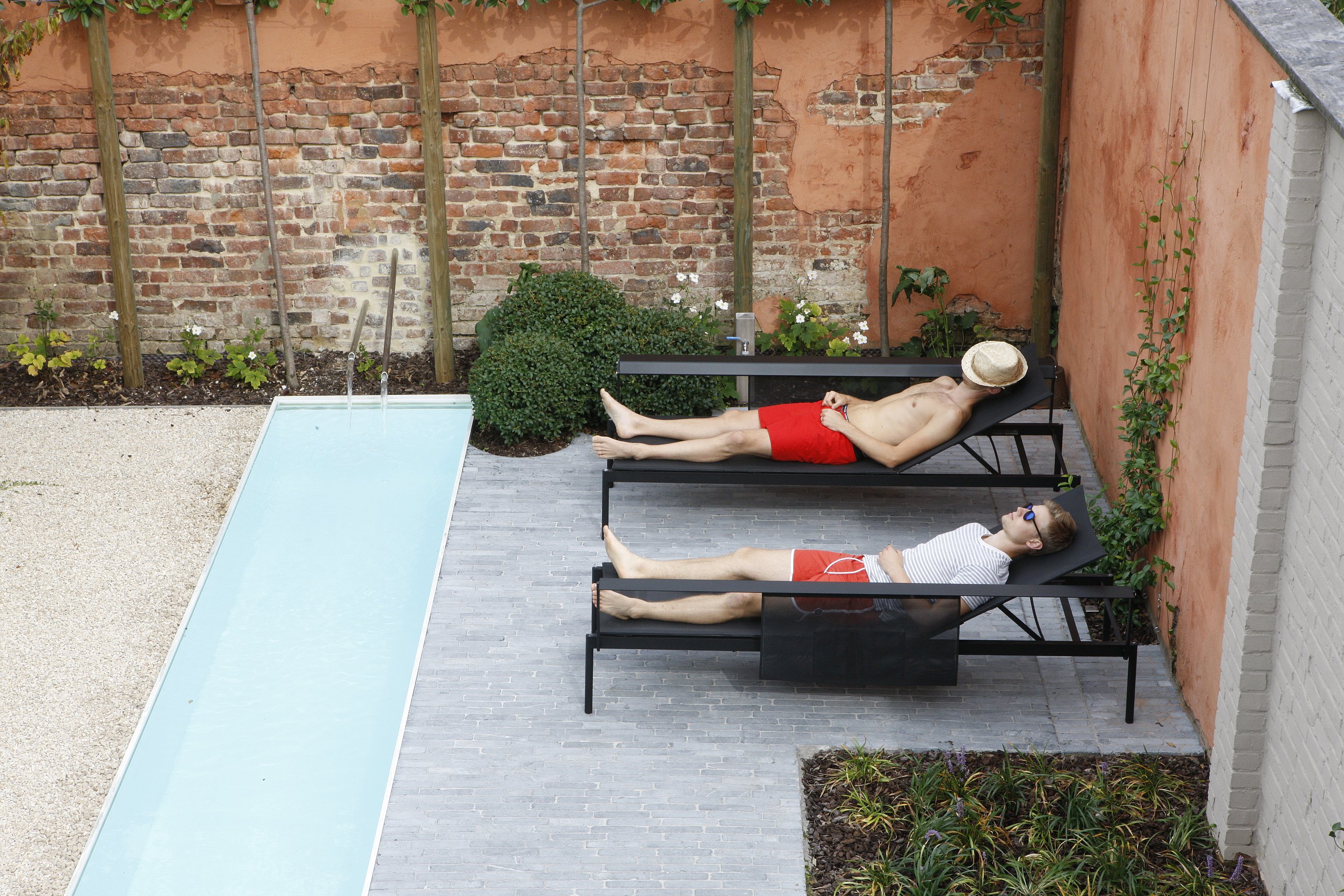 Draufsicht auf zwei Personen, die sich auf schwarzen Liegestühlen neben einem langen, schmalen Pool in einem abgeschiedenen Innenhof mit Backstein- und Putzwänden sonnen.