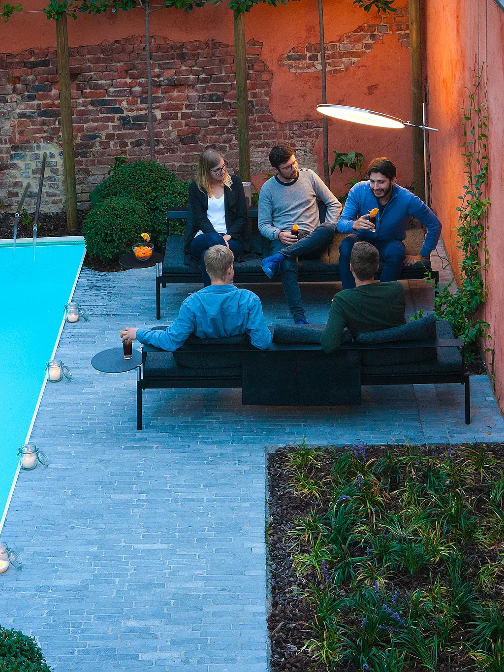 Cinq amis se détendent sur des chaises longues noires d'extérieur, au bord d'une piscine étroite dans une cour privée entourée de murs en briques, au crépuscule, sous un parasol rond lumineux.