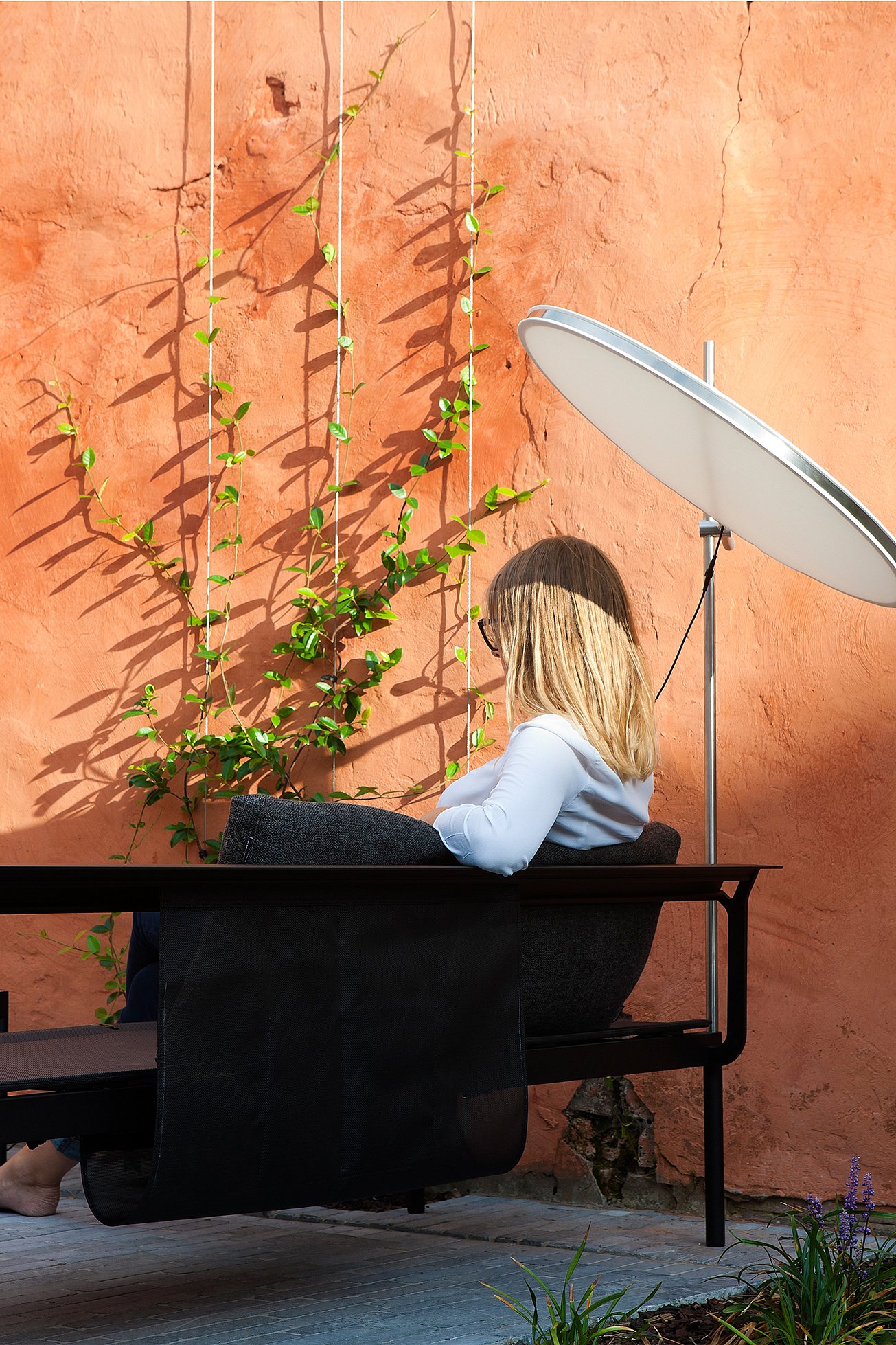 Eine Frau sitzt auf einer schwarzen Gartenliege neben einem runden weißen Sonnenschirm und blickt auf eine Kletterpflanze vor einer warmen Terrakotta-Wand.