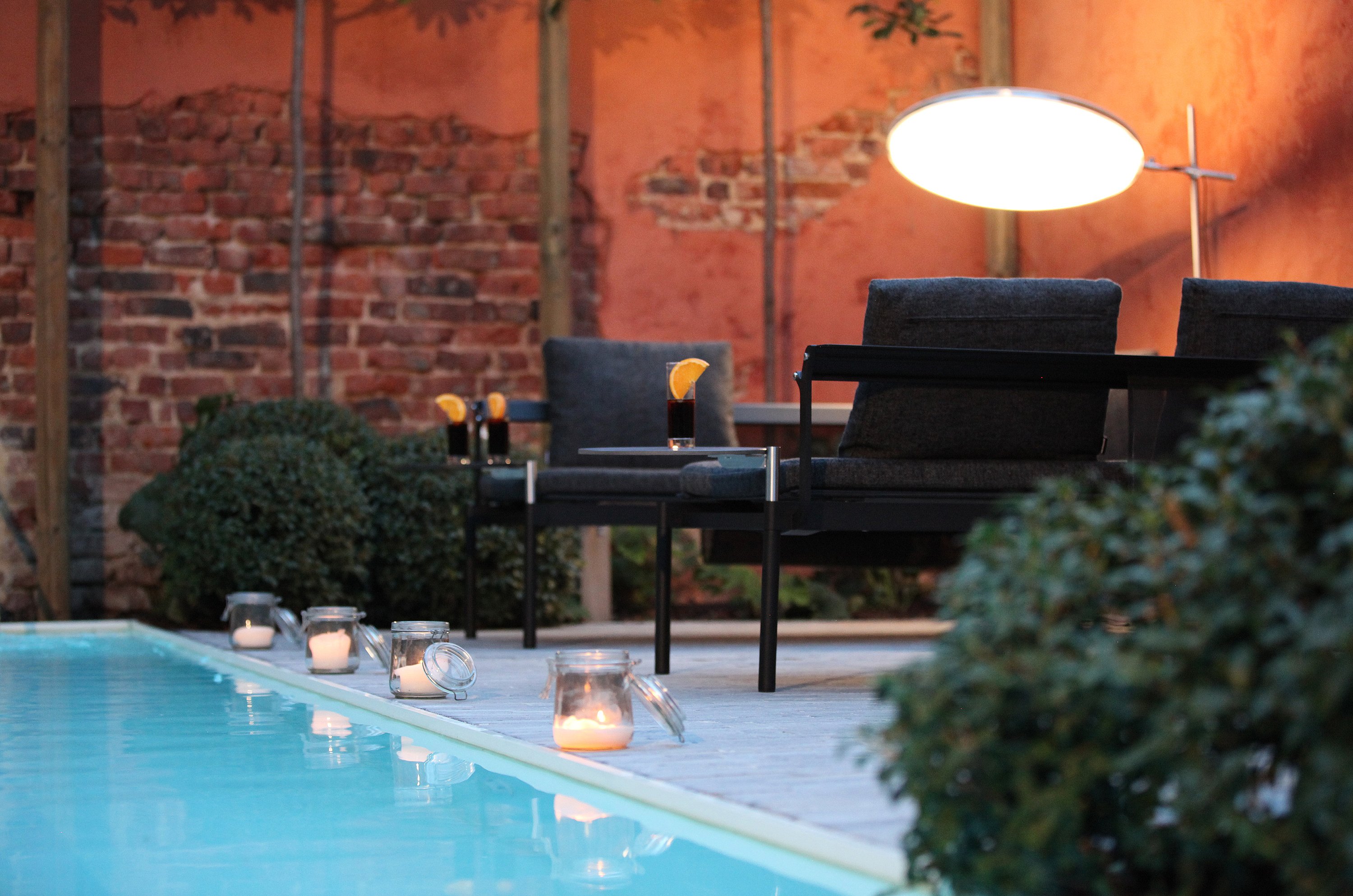   Abendliche Poolterrasse mit schwarzen Liegestühlen, die wie Sofas angeordnet sind, einer leuchtenden runden Lampe darüber, Kerzen am Wasserrand und Getränken auf Beistelltischen.