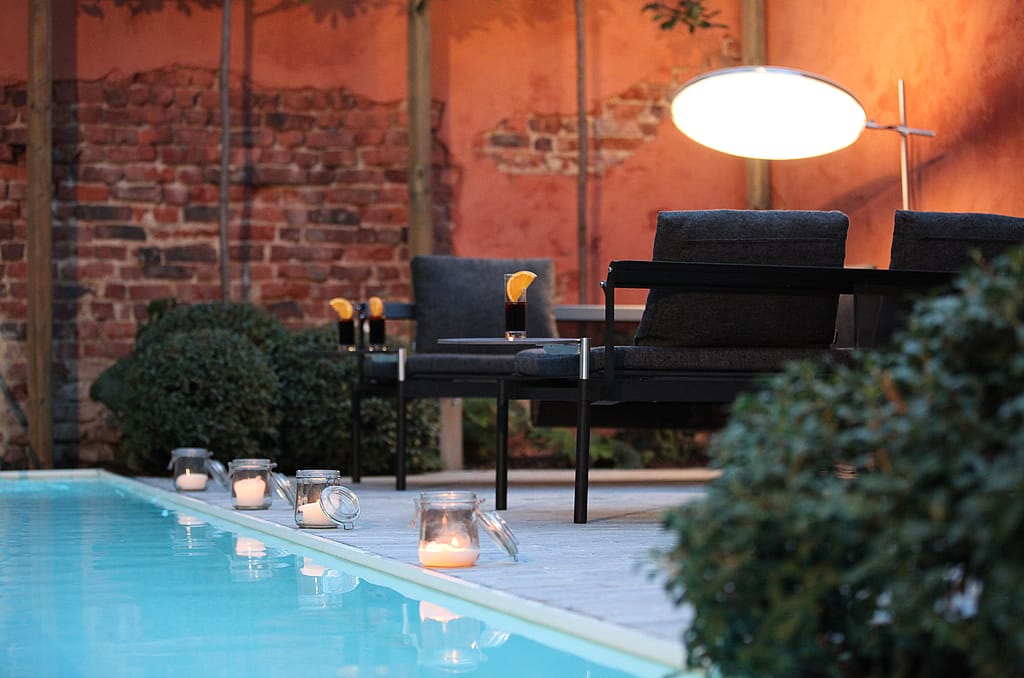 Abendliche Poolterrasse mit schwarzen Liegestühlen, die wie Sofas angeordnet sind, einer leuchtenden runden Lampe darüber, Kerzen am Wasserrand und Getränken auf Beistelltischen.