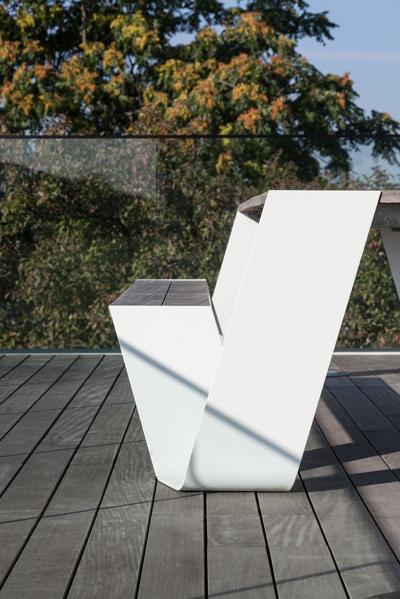 Side profile of a white Hopper picnic table with wooden seat and tabletop on a balcony terrace, with trees visible beyond the glass balustrade.