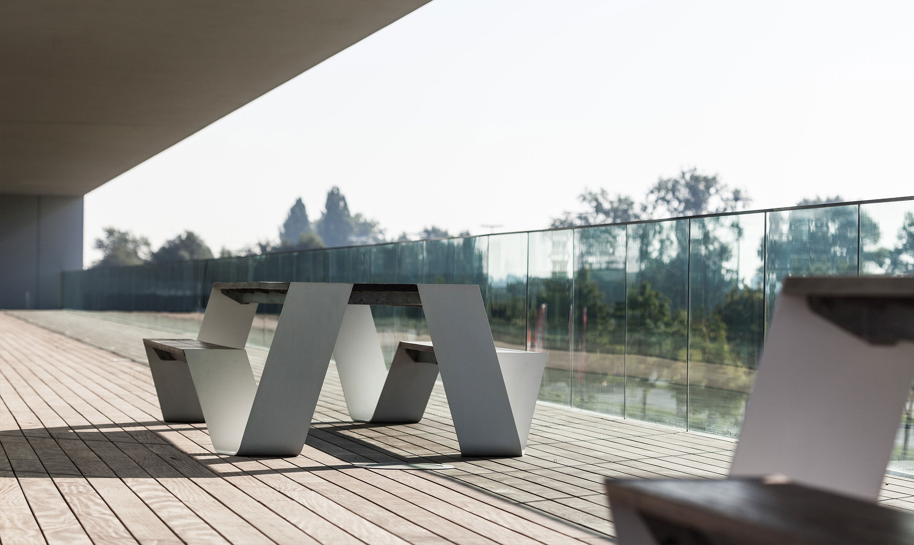 Table de pique-nique Single Hopper placée sur un long balcon abrité avec garde-corps en verre, terrasse ensoleillée et verdure douce au loin.