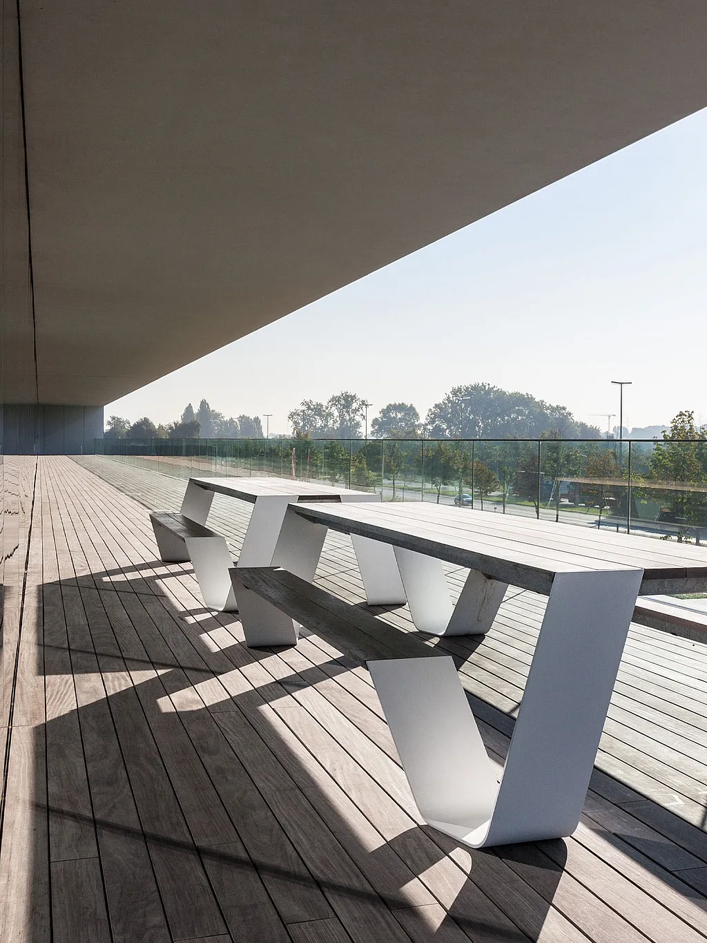 Long covered office balcony with a white Hopper picnic table on dark wooden decking, mirrored in the glass façade and bordered by a transparent glass railing.