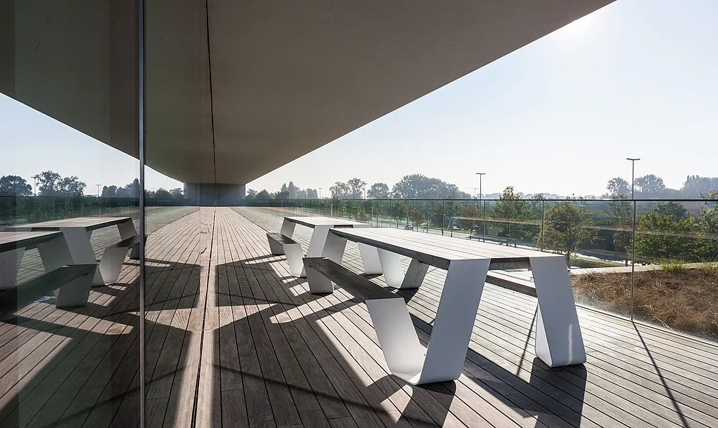 Lang overdekt kantoorbalkon met een witte Hopper picknicktafel op donker houten terras, gespiegeld in de glazen gevel en begrensd door een transparante glazen reling.