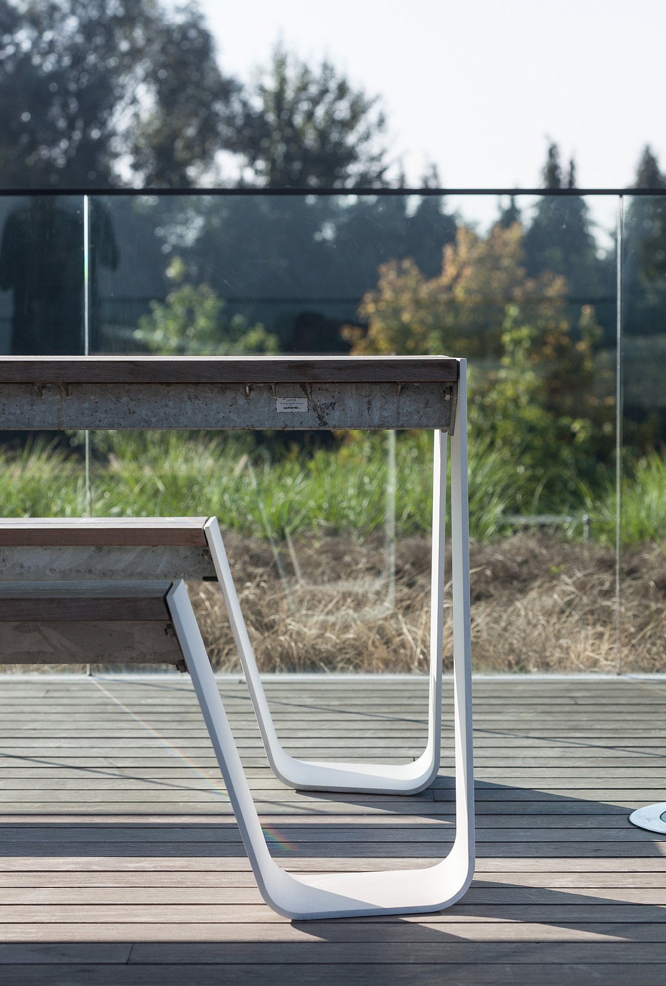 Gros plan sur les pieds incurvés fins et blancs de la table Hopper et le banc en bois sombre sur une terrasse de style toit avec des plantations au-delà du bord du verre.