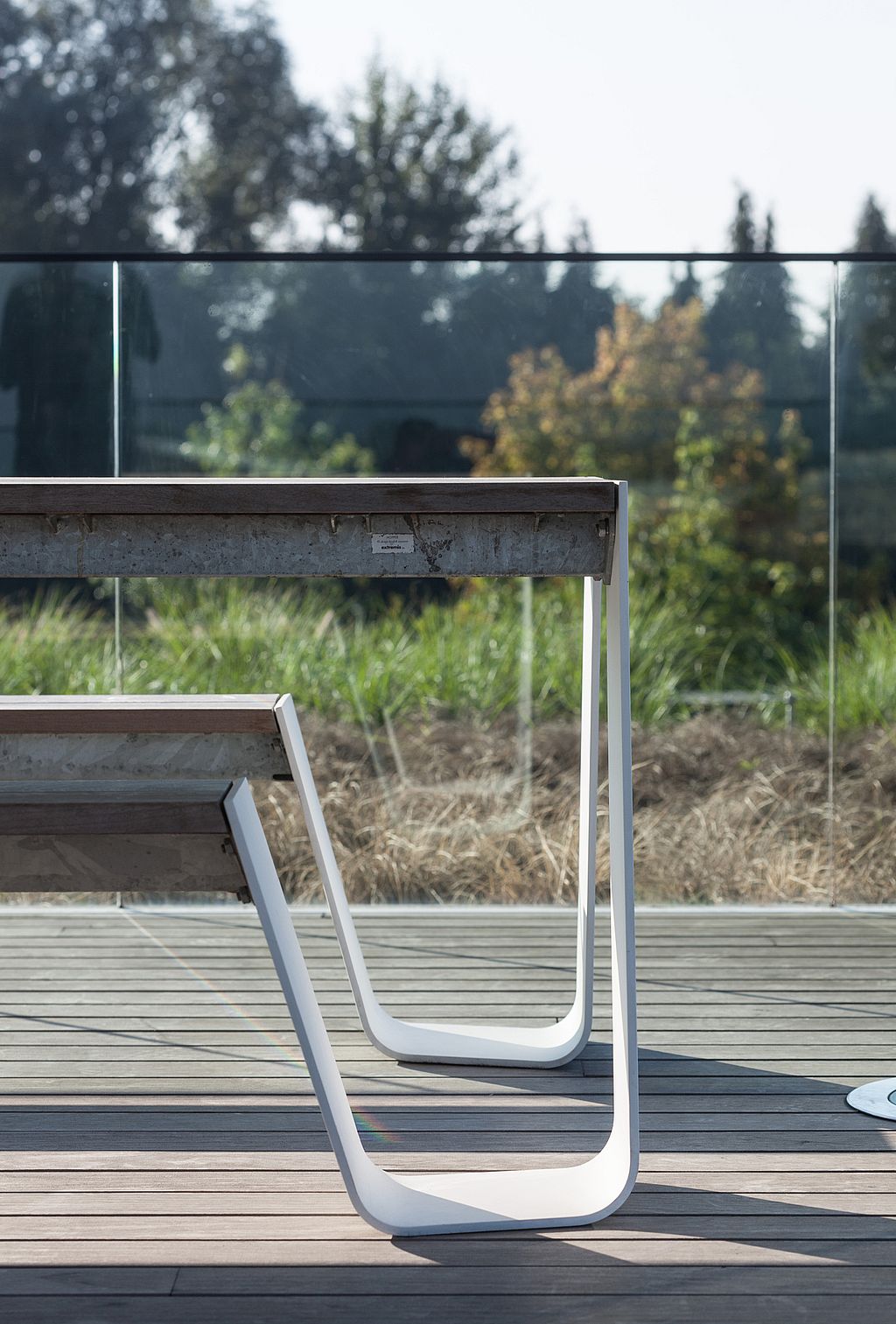 Gros plan sur les pieds incurvés fins et blancs de la table Hopper et le banc en bois sombre sur une terrasse de style toit avec des plantations au-delà du bord du verre.