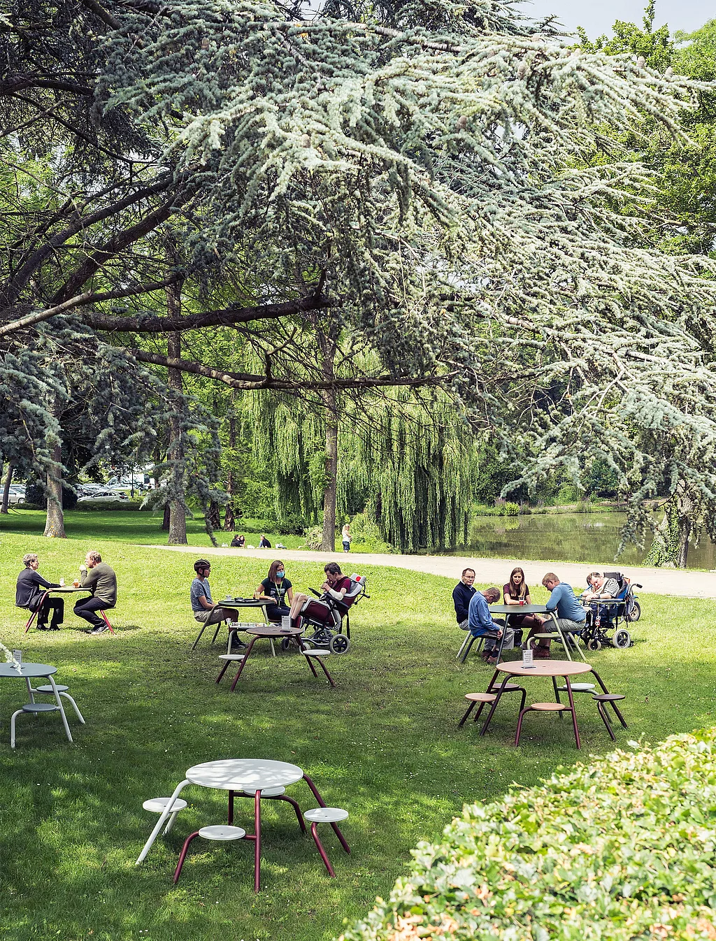 Menschen versammeln sich an farbenfrohen, runden Virus-Picknicktischen, die auf einer grünen Parkwiese unter großen, überhängenden Bäumen verteilt sind. Im Hintergrund befinden sich ein Teich und ein Spazierweg.