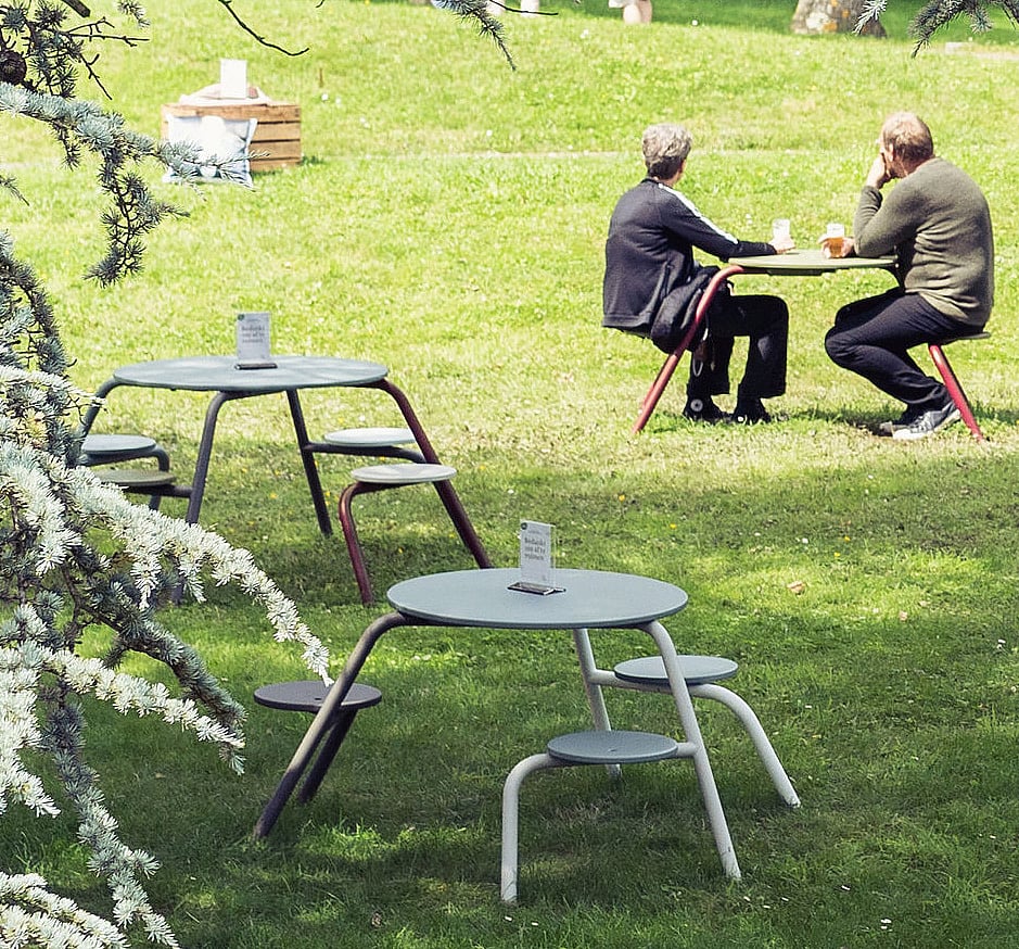   Zwei leere Virus-Tische mit runden Tischplatten und Hockern stehen auf dem Rasen. Zwei Personen unterhalten sich an einem anderen Tisch im sonnigen Park dahinter.