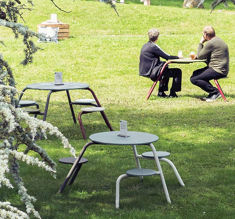 Zwei leere Virus-Tische mit runden Tischplatten und Hockern stehen auf dem Rasen. Zwei Personen unterhalten sich an einem anderen Tisch im sonnigen Park dahinter.