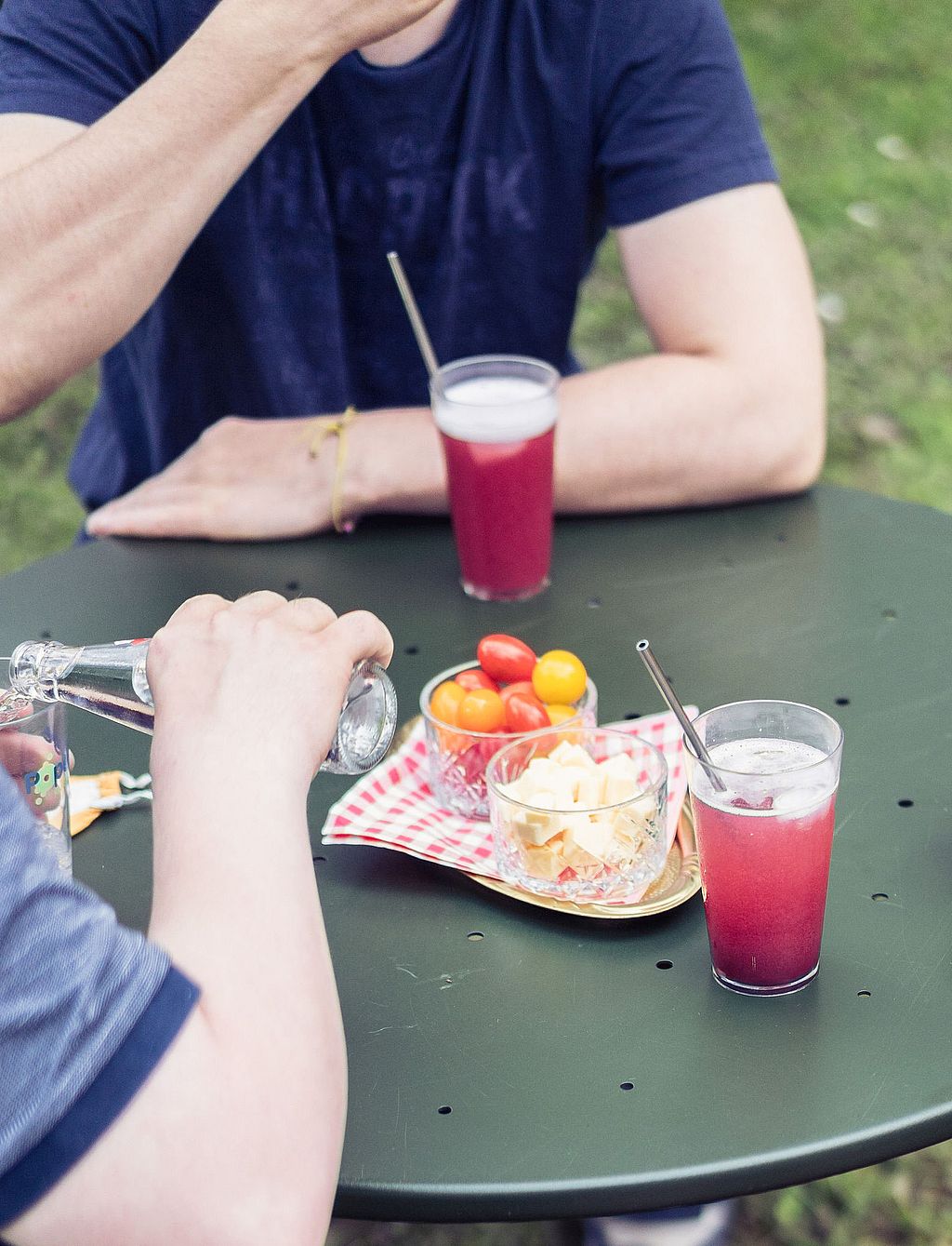 Nahaufnahme: Snacks und leuchtend pinkfarbene Getränke auf einer dunkelgrünen Virus-Tischplatte. Menschen sitzen draußen darum herum.