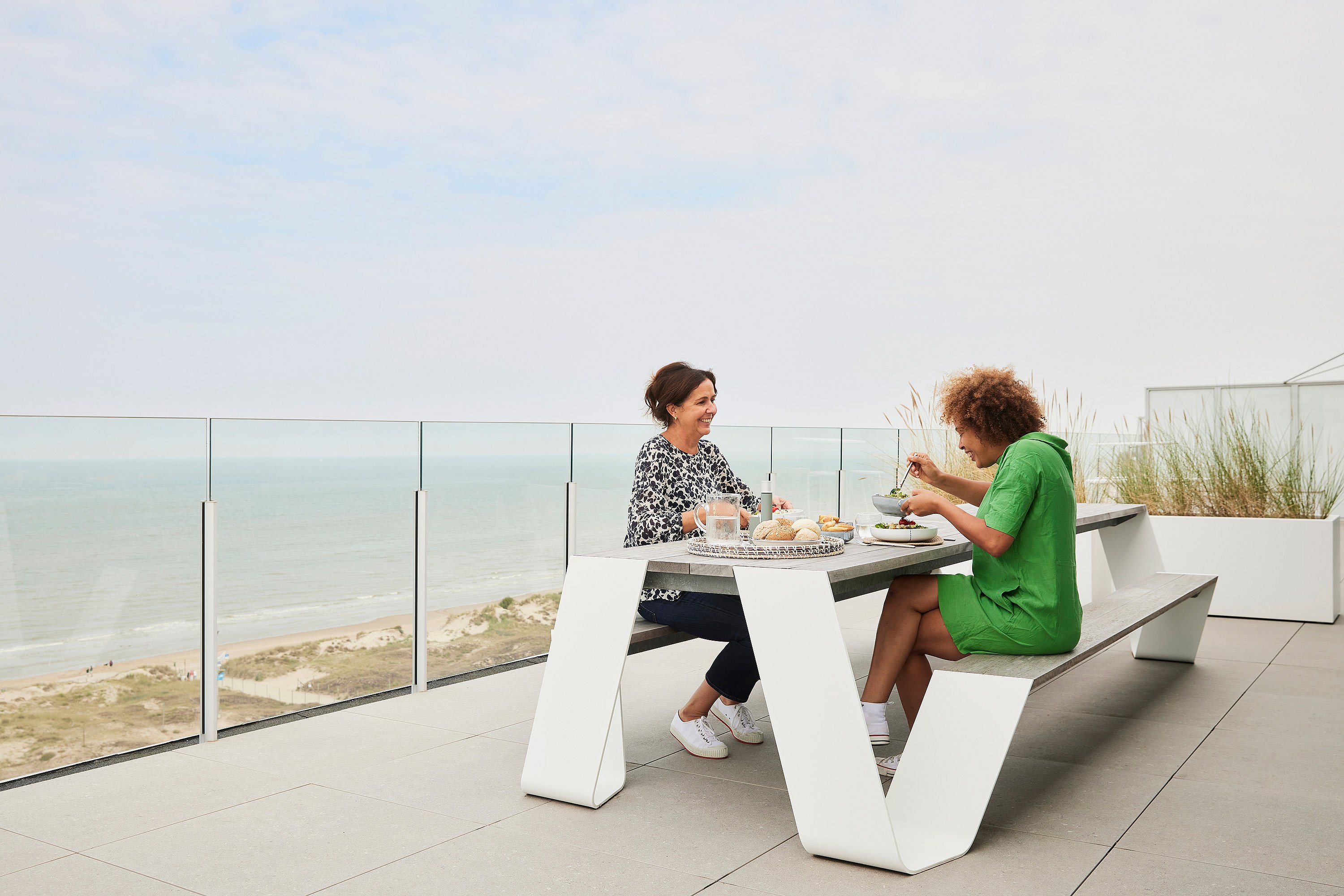 Two women enjoy lunch at a white Hopper picnic table on a rooftop terrace with glass railings and sweeping views of the North Sea and dunes.